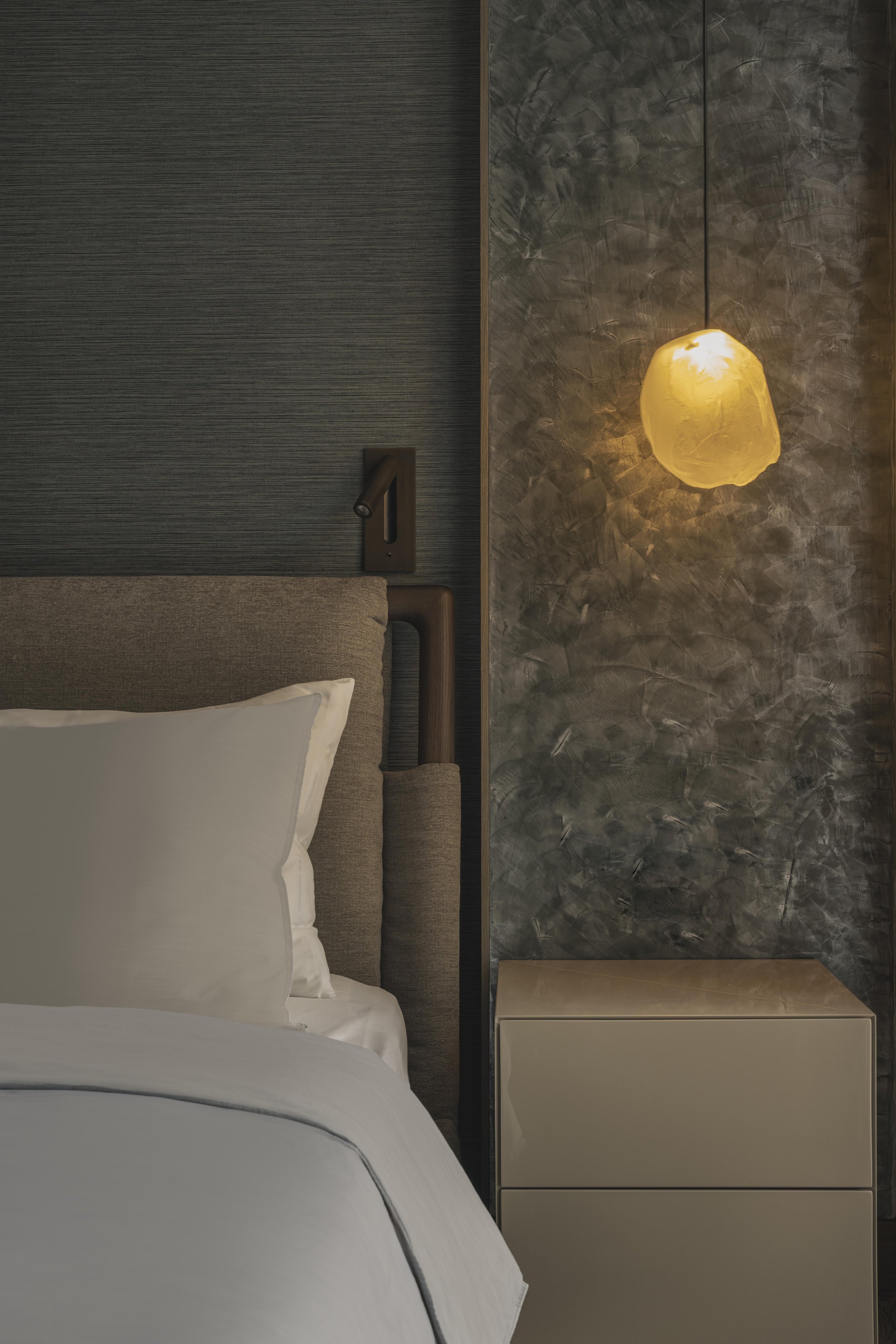 Interior of a modern bedroom with a bed, bedside table, textured grey walls, a decorative wall lamp, and a marble accent wall.