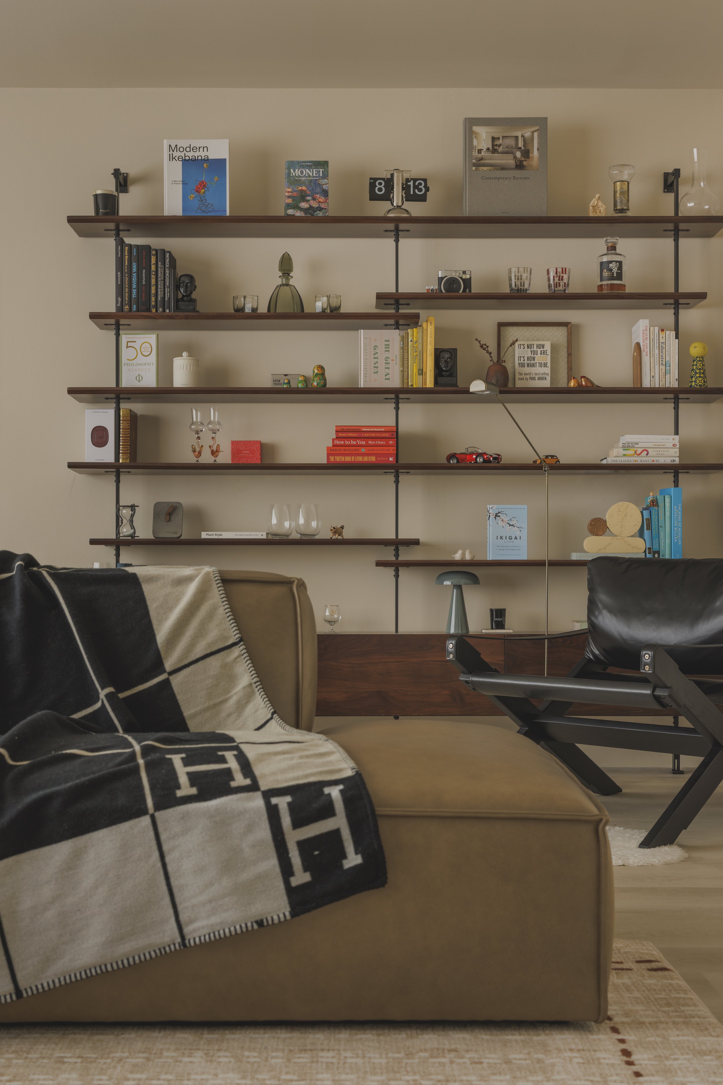 Living room with beige sofa, black and white checkered throw blanket, a wooden wall shelf with books, decorative items, and framed artwork, beige rug, and part of a black lounge chair visible.