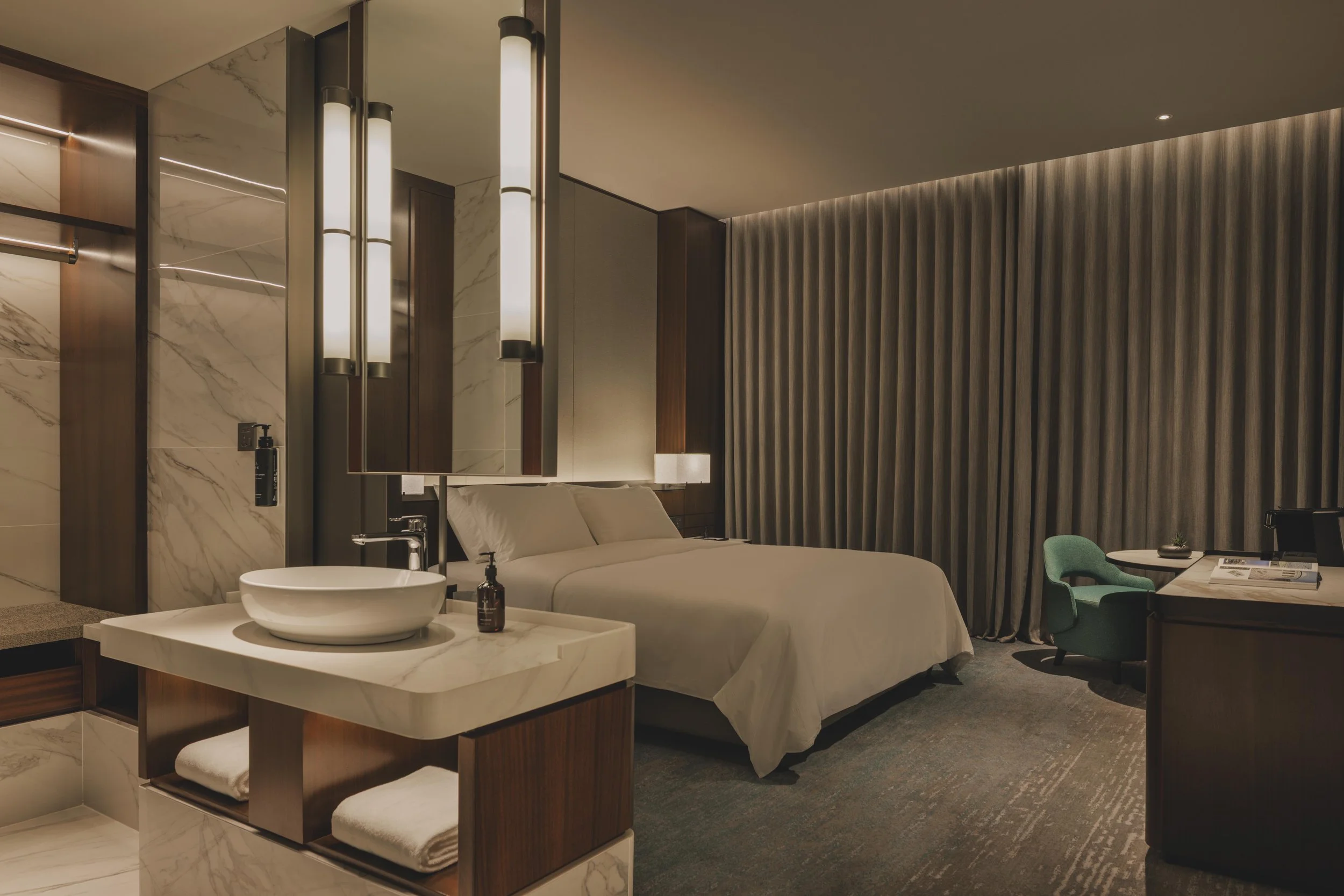 Modern hotel room with a bed, a mirror with vertical lights, a marble-topped vanity with a bowl sink, a bedside table, a small desk with a green chair, and textured wall paneling.