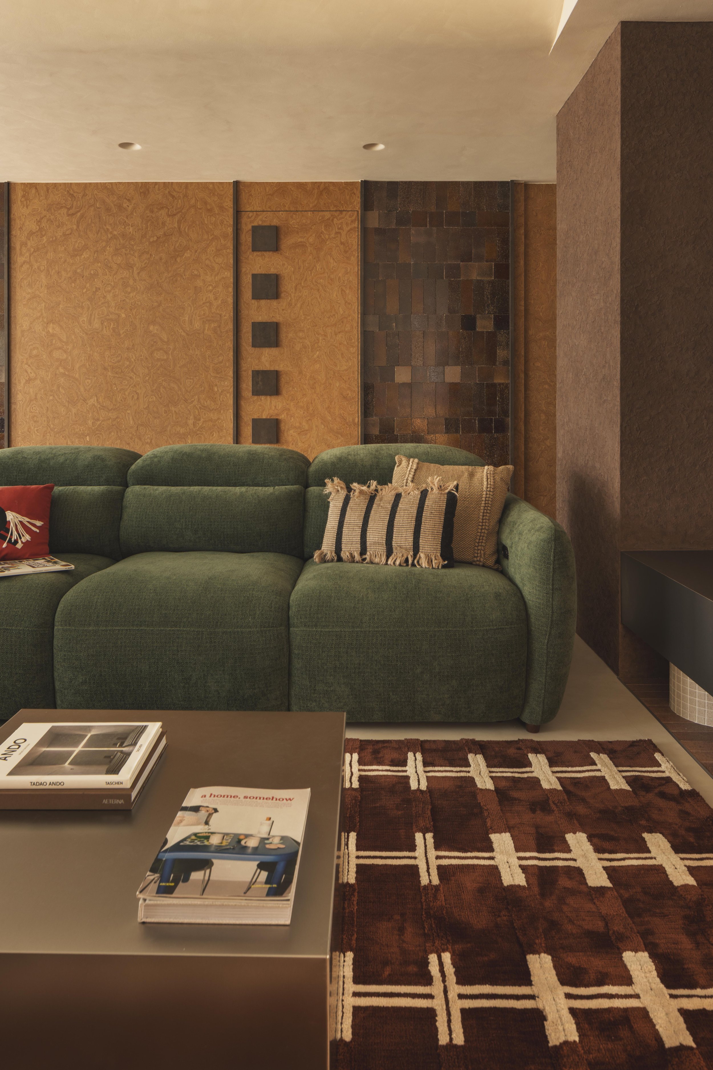 Living room with green sofa, brown patterned area rug, brown coffee table with books and magazines, textured wall with wood and stone paneling, pillows on the sofa, small table on the right side.