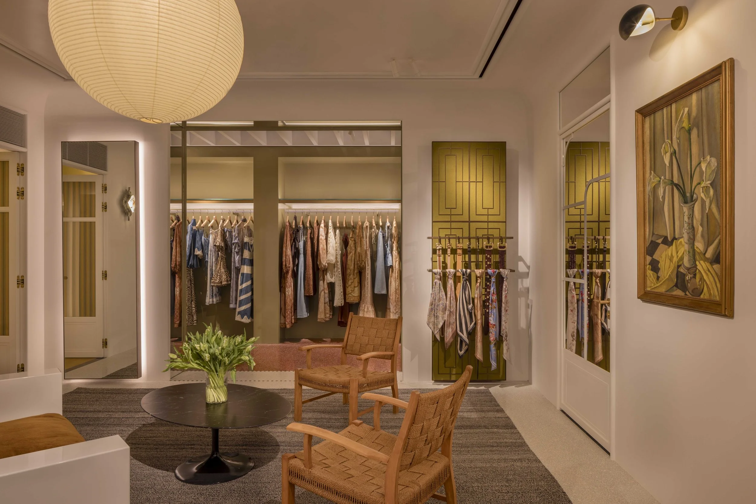 Interior of a clothing boutique with racks of dresses and suits, a round table with a vase of plants, and chairs. The walls have artwork and a mirror, with warm lighting.