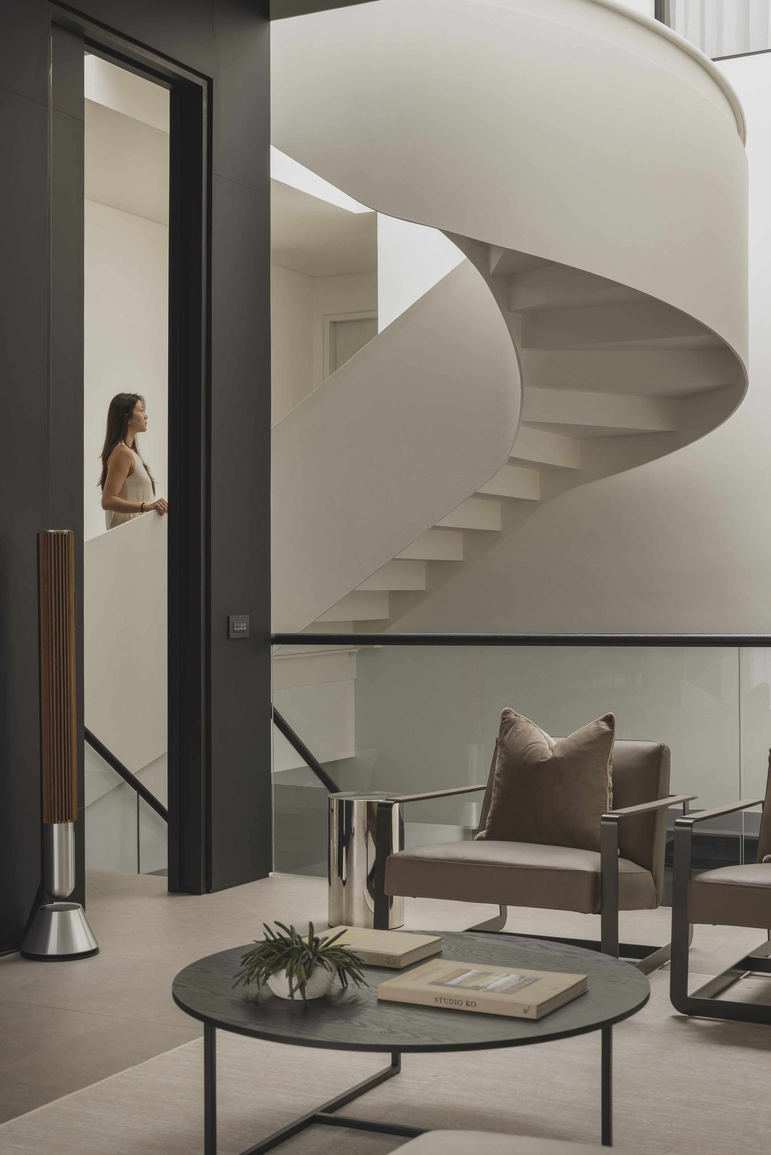 Interior view of a modern, minimalist living space with a curved white staircase, beige armchairs with pillows, a round coffee table with books and a plant, and an elevator with a woman standing near it, seen through a glass wall.