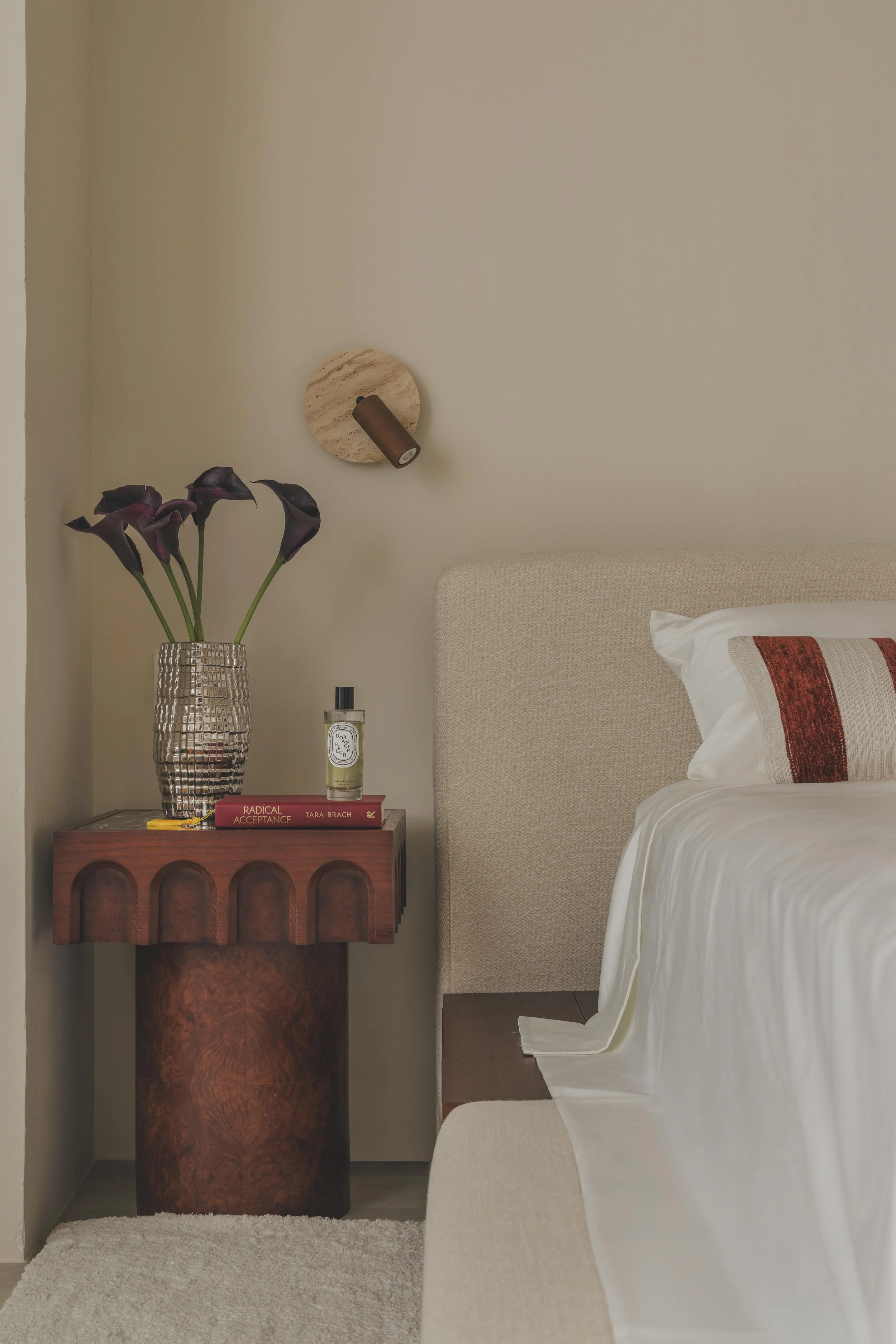 A beige headboard next to a cream-colored textured wall with a wooden wall-mounted light fixture. A bedside table with a glass vase of dark purple calla lilies, a bottle of candle, and two books, one titled 'Radical Acceptance' and the other with a red cover, is beside the bed. The bed has white bedding with a red and beige decorative pillow. The floor is light-colored wood, and there is a cream-colored rug in front of the bedside table.