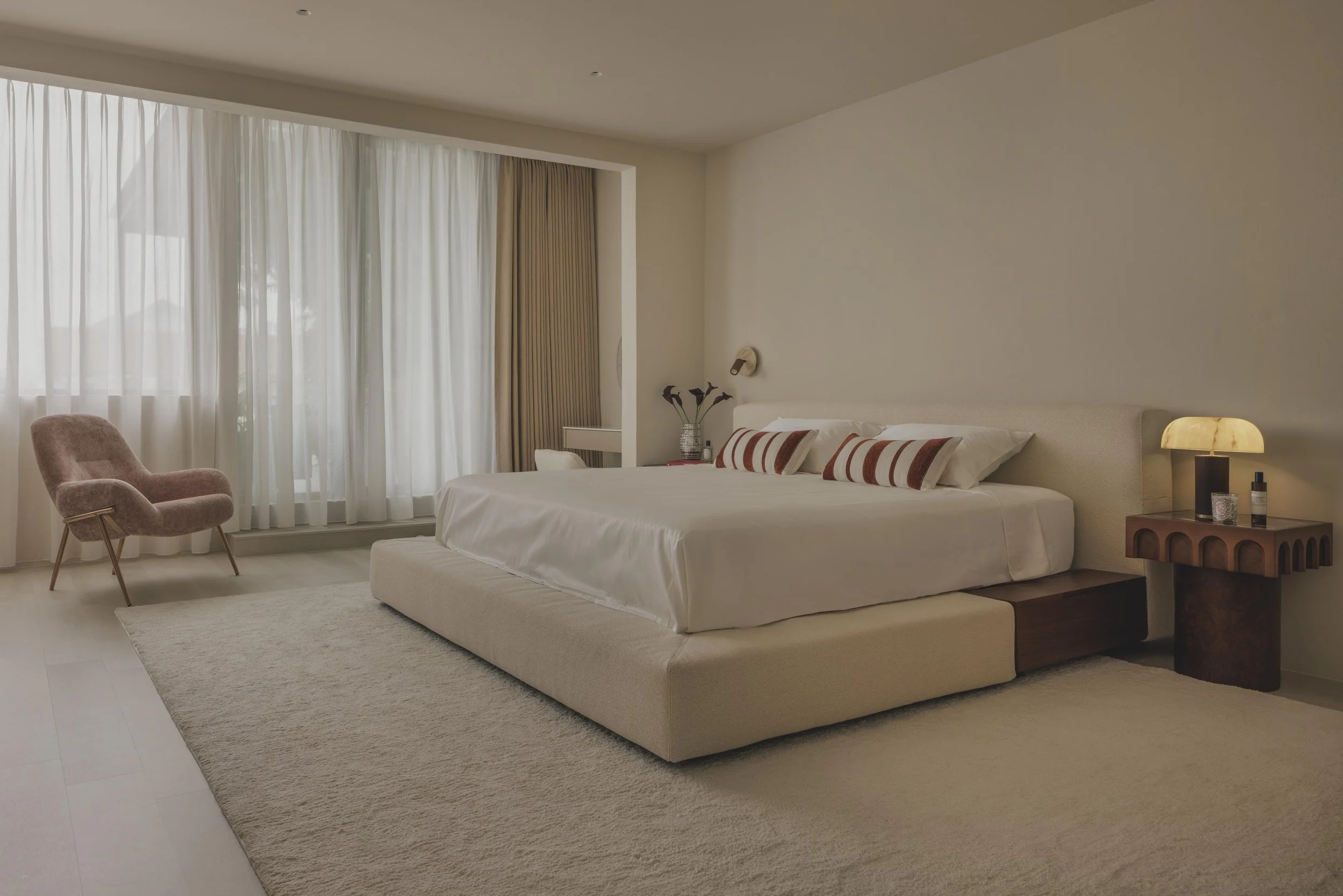 Modern bedroom with a large bed, pink armchair, side table with lamp and flowers, and sheer curtains that let in natural light.