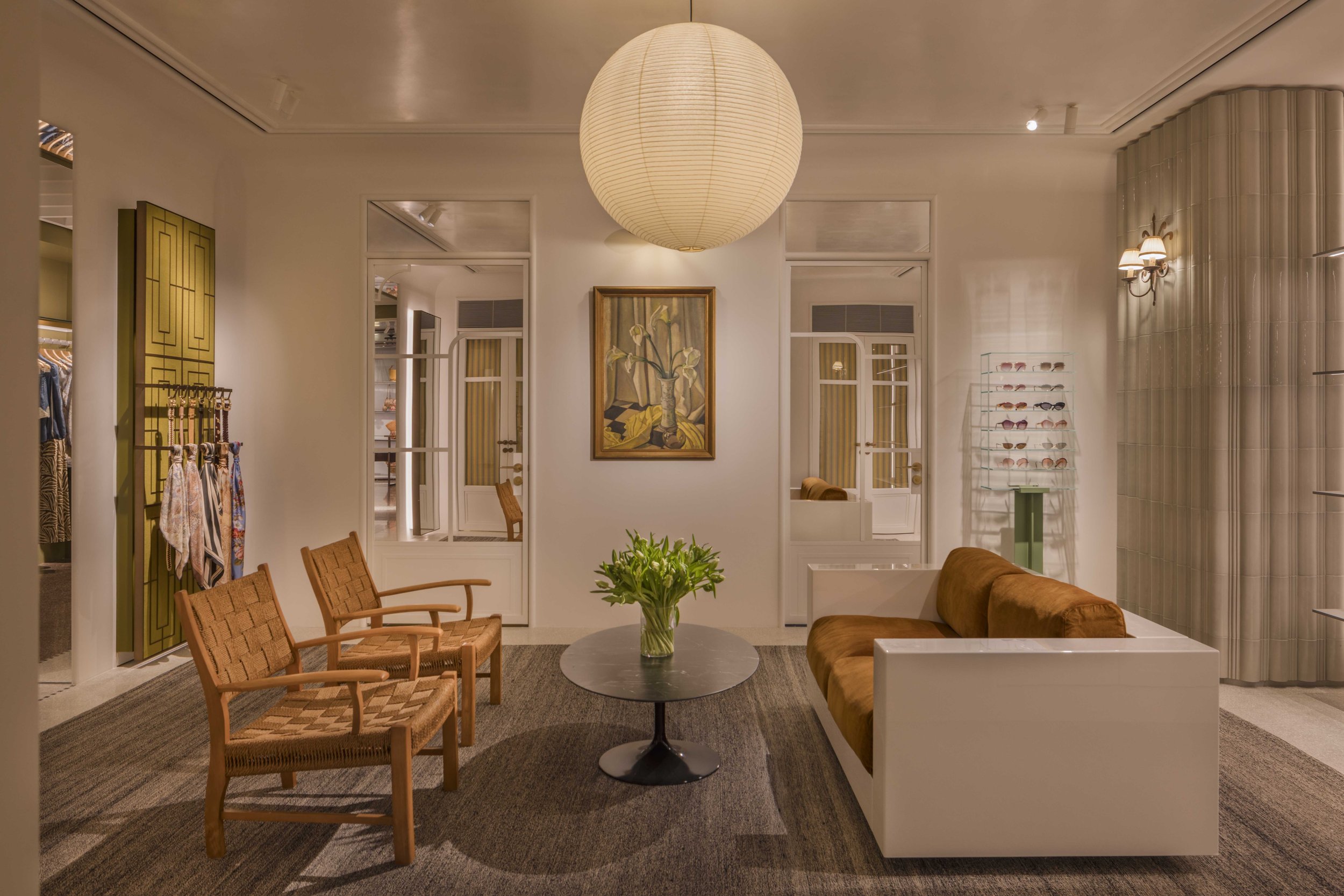 A waiting area inside a retail store with wooden chairs, a round marble table with a vase of green plants, a brown couch, art on the wall, a large paper lantern hanging from the ceiling, and a display of sunglasses on a stand.
