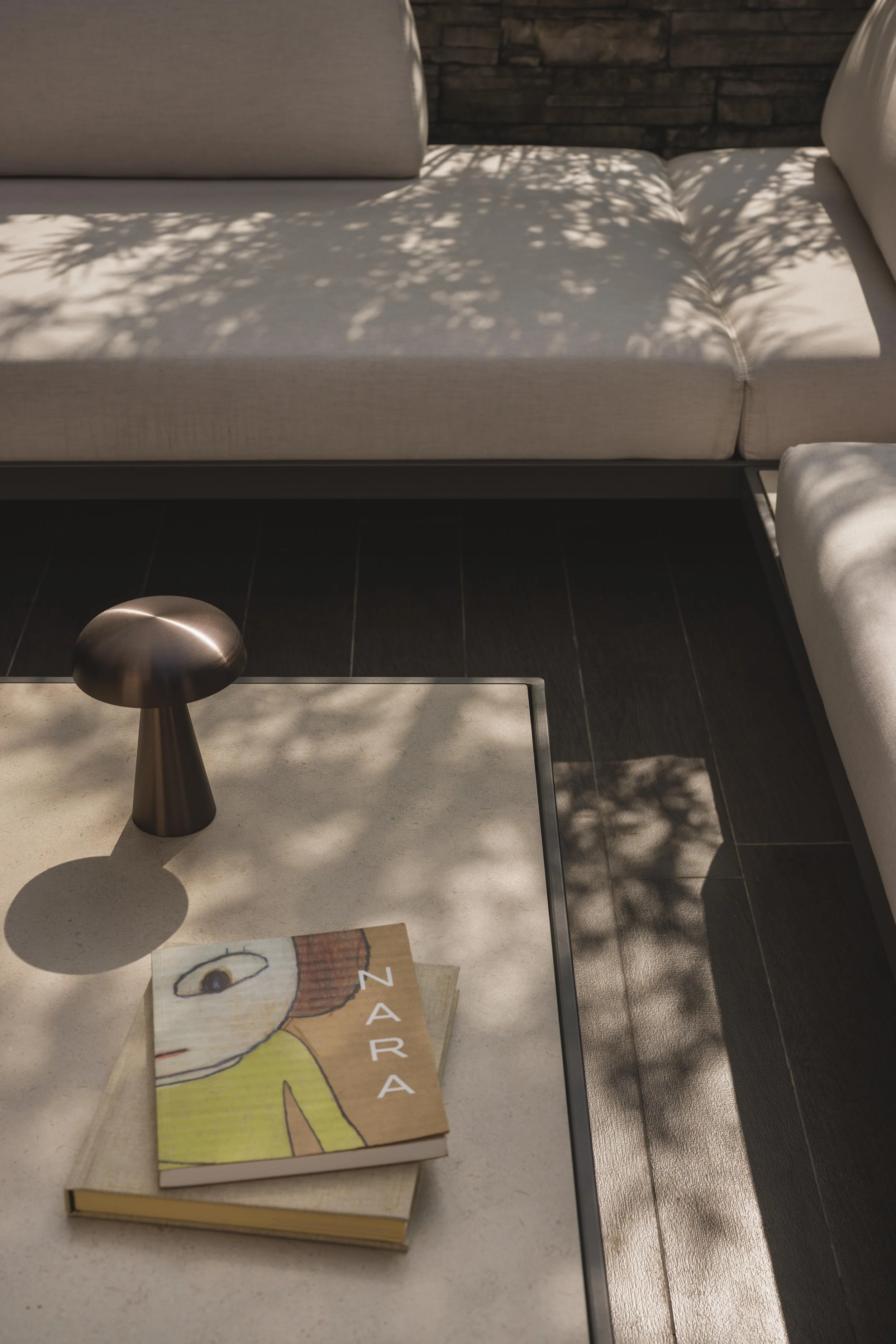A beige-colored book on a white table with a drawing of a person on the cover. The drawing depicts a person with brown hair, wearing a yellow shirt. A black pencil is lying on top of the book, casting a shadow. There are some blurred items beneath the book, and a brown object on the edge of the table. The background shows a sofa with white cushions, a dark wood floor, and shaded sunlight.