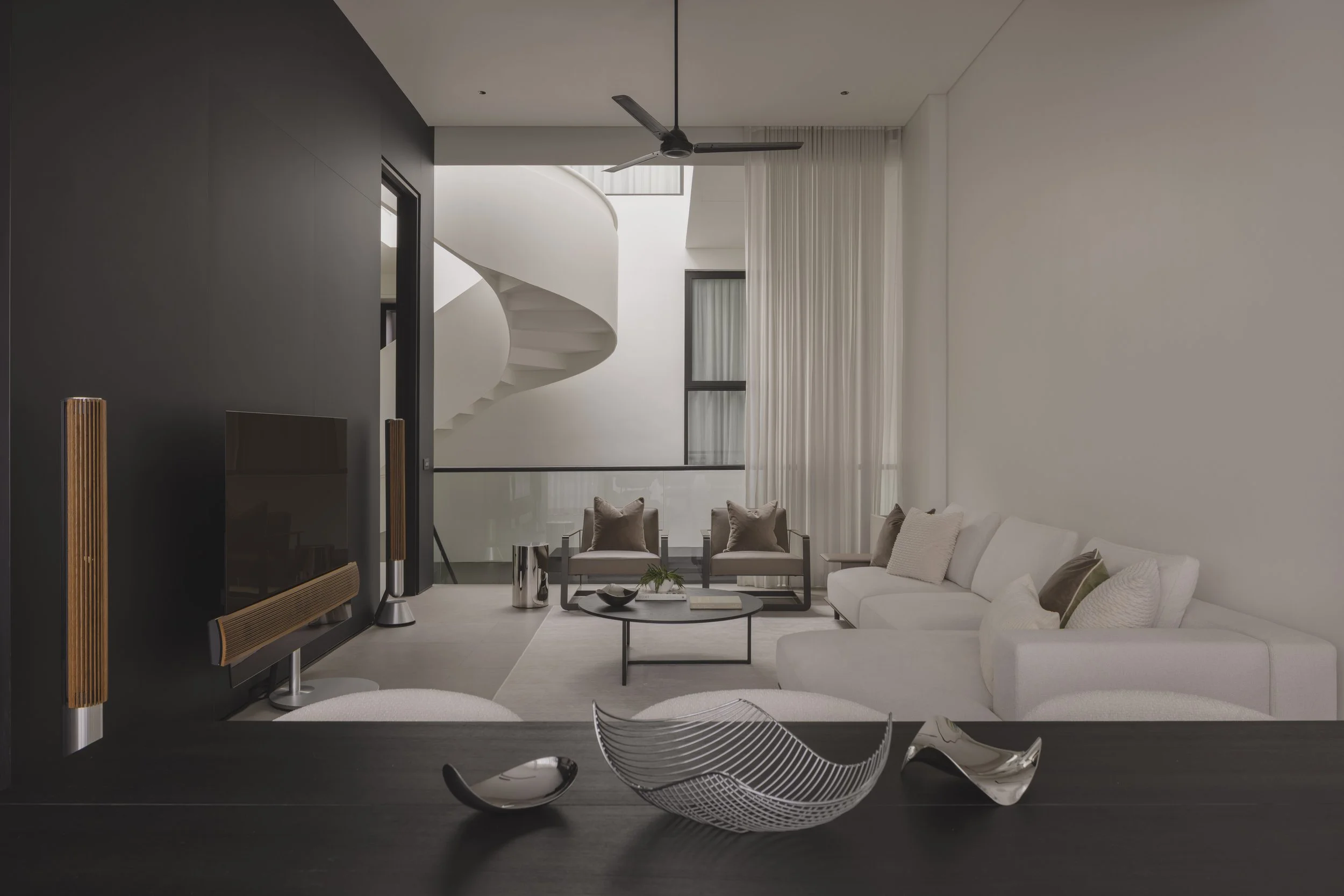 Modern living room with white sofa, black coffee table, and contemporary chairs in front of a large window with sheer curtains, spiral staircase, and ceiling fan.