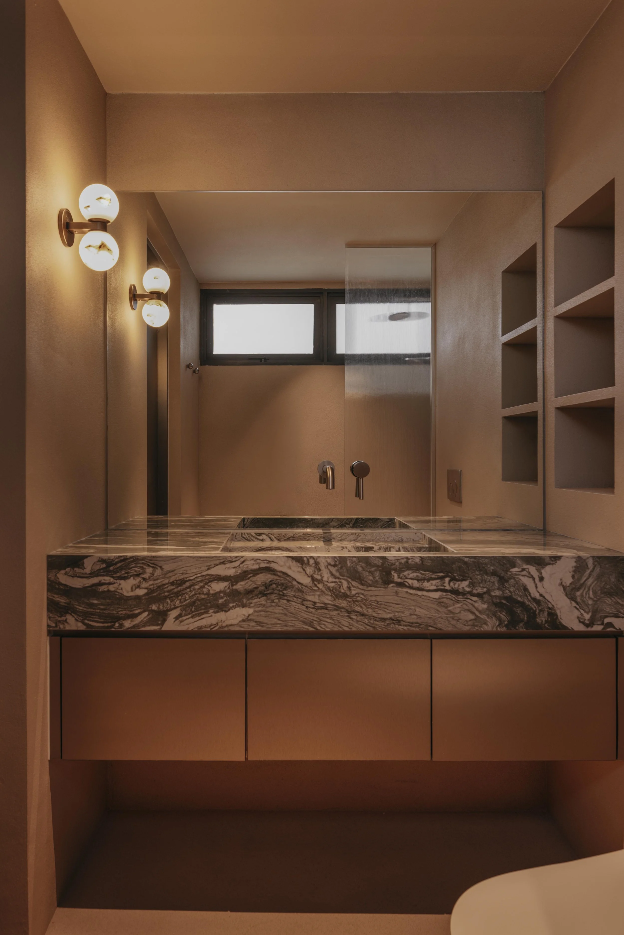 Bathroom with beige walls, three round bubble style wall lights, a rectangular window, a marble countertop with a built-in sink, and open shelving on the right side.