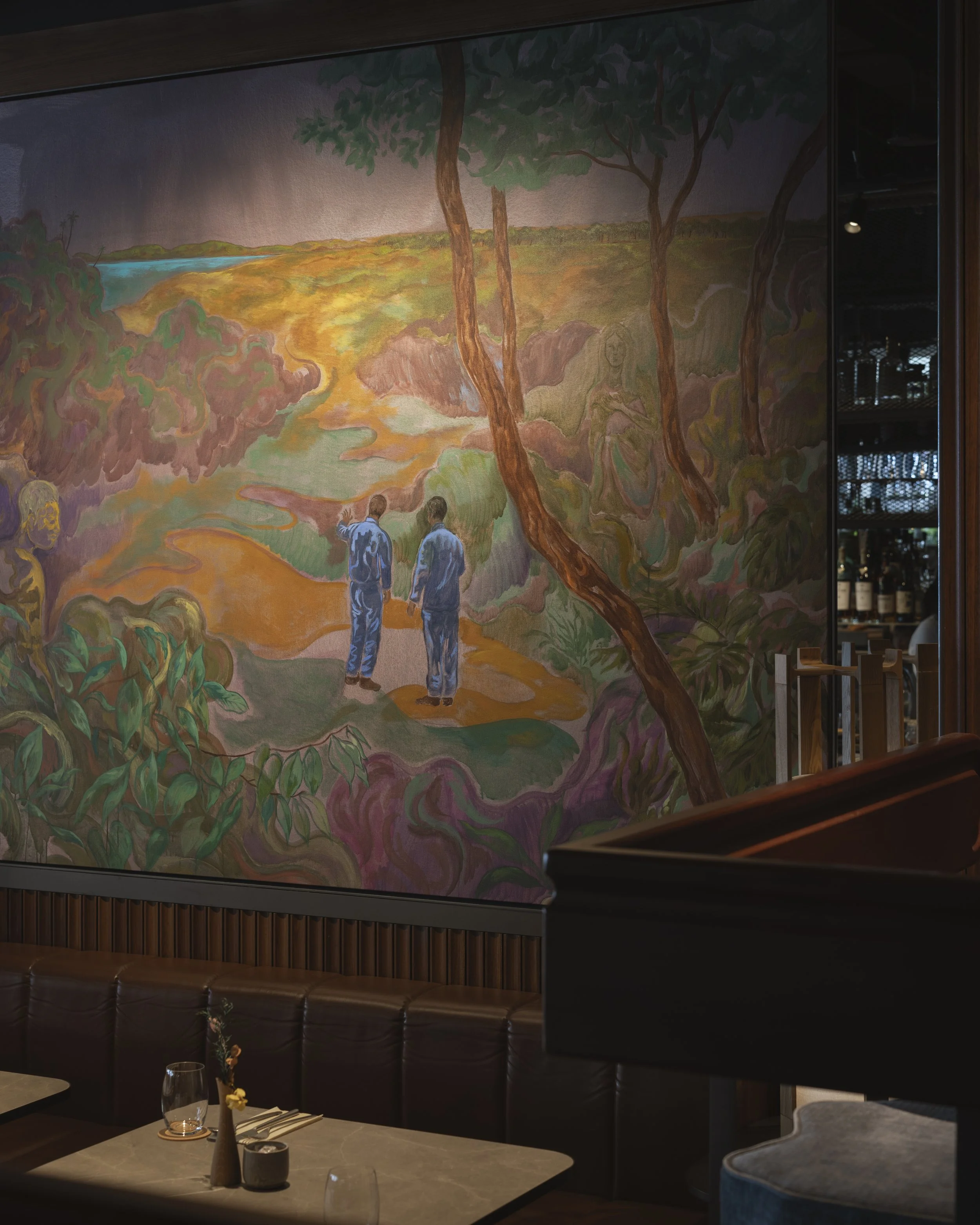 Interior of a restaurant featuring a large mural painting on the wall depicting a landscape with trees, a river, and two people walking on a path, with a table set with glasses, a vase with flowers, and tableware in the foreground.