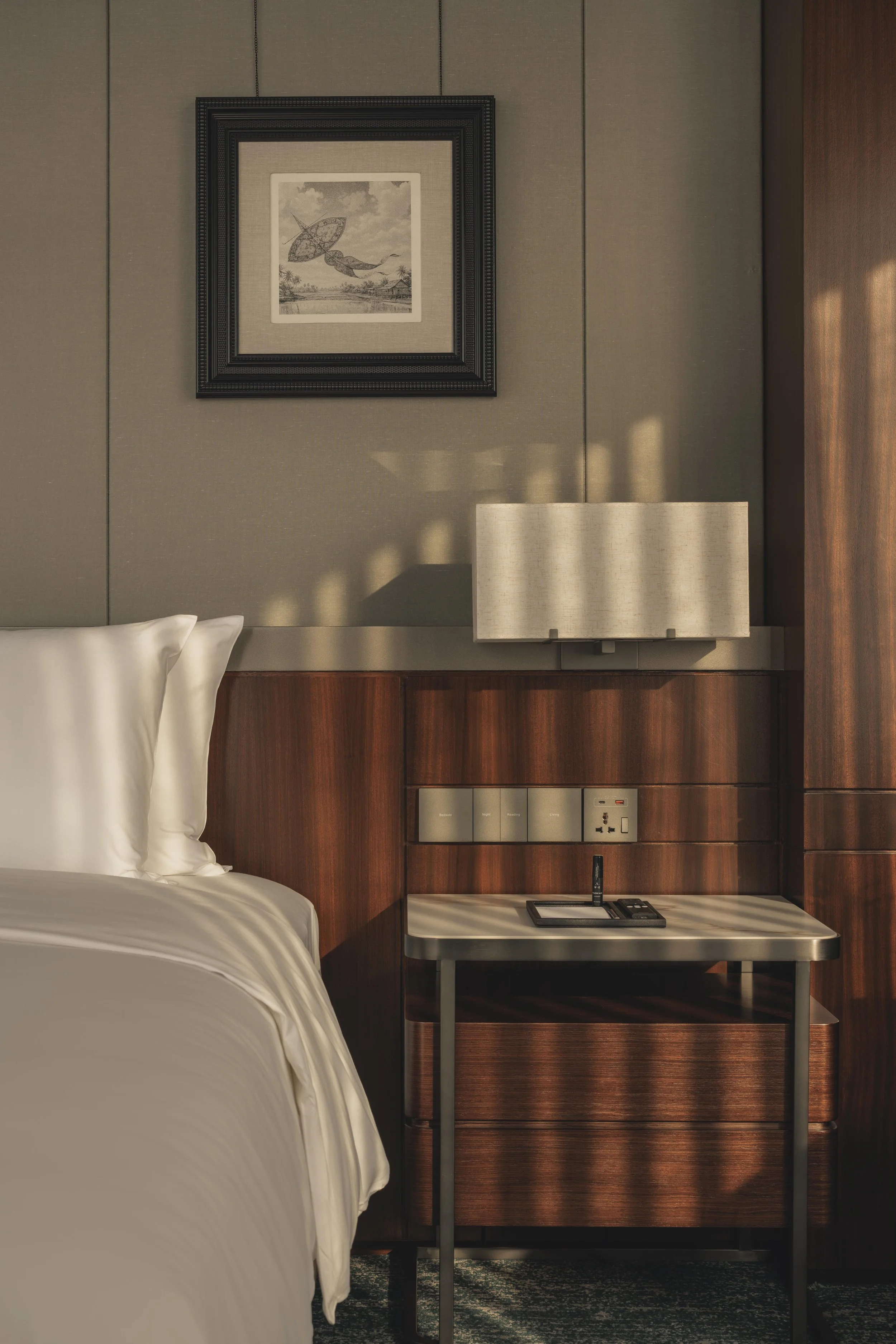 A hotel room interior featuring a bed with white linens, a bedside table with a remote control, a pen, and a paper, a wall-mounted lamp, framed artwork depicting a hot air balloon and a whale, and a power outlet with a plug.