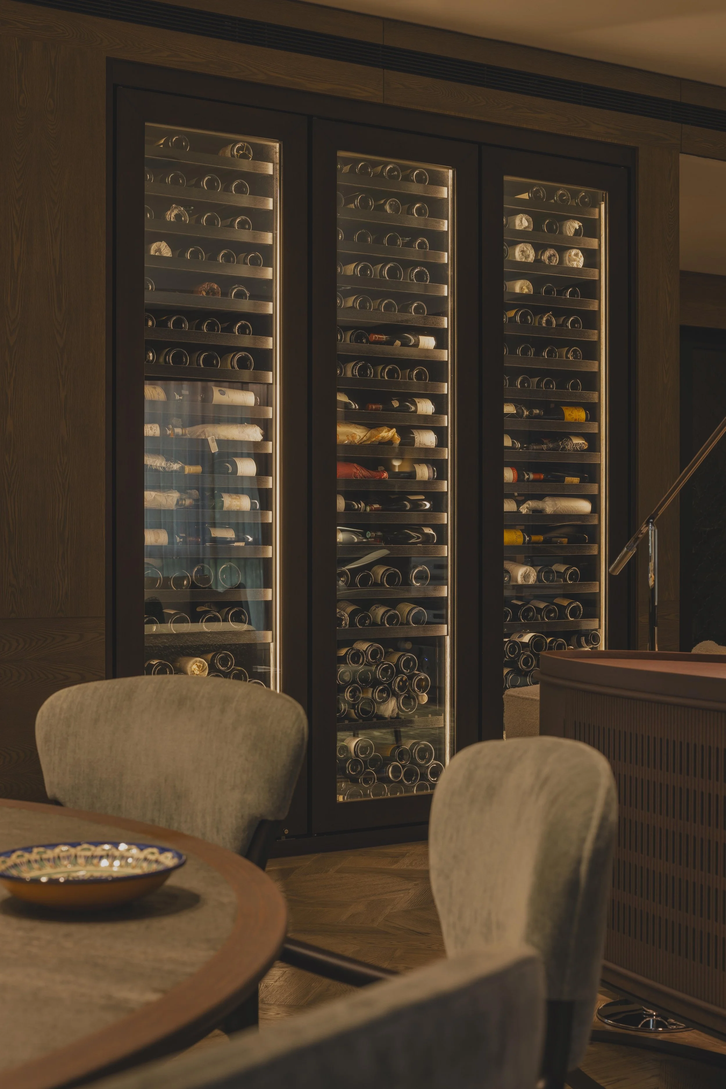 A wine storage unit with glass doors, filled with bottles of wine, placed in a modern, warmly lit room with wooden accents. Part of a dining area with chairs and a table visible in the foreground.