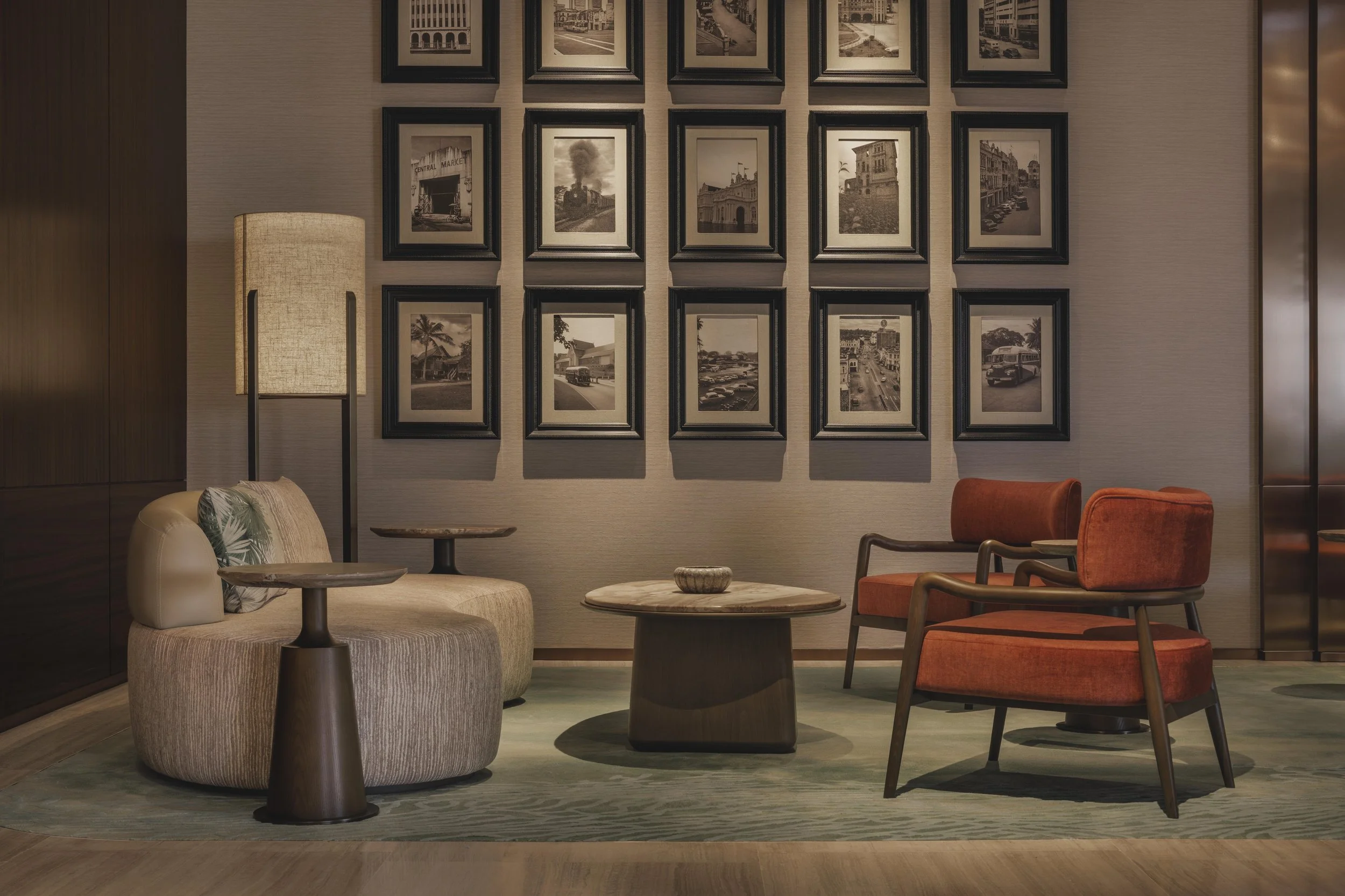 A cozy hotel lobby with a collection of black and white framed photographs on a beige wall. There is a beige upholstered armchair with a patterned pillow, a small wooden side table, and a modern floor lamp with a textured shade. Also, there are two orange velvet chairs and a marble-topped coffee table with a decorative bowl.