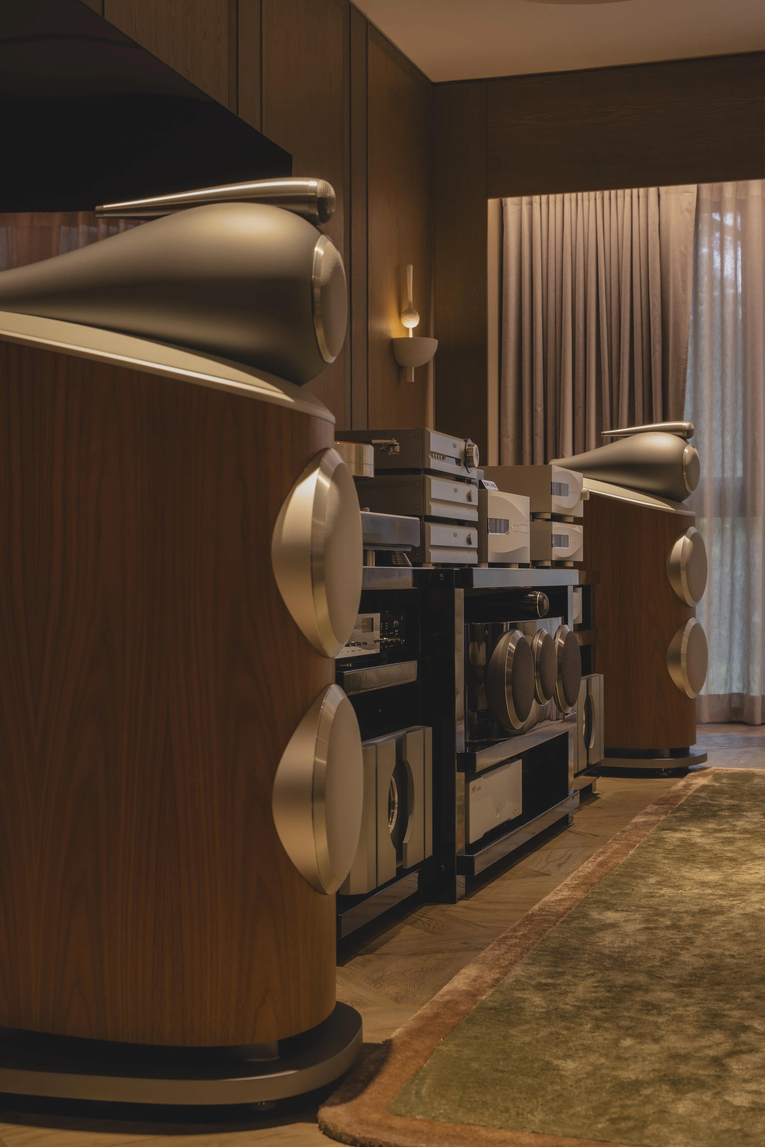 A home theater with wooden paneling on the walls, large speakers embedded in a curved wooden cabinet, and a vintage stereo system on a black stand. A curtain covers a window, and a beige and green area rug is on the wooden floor.