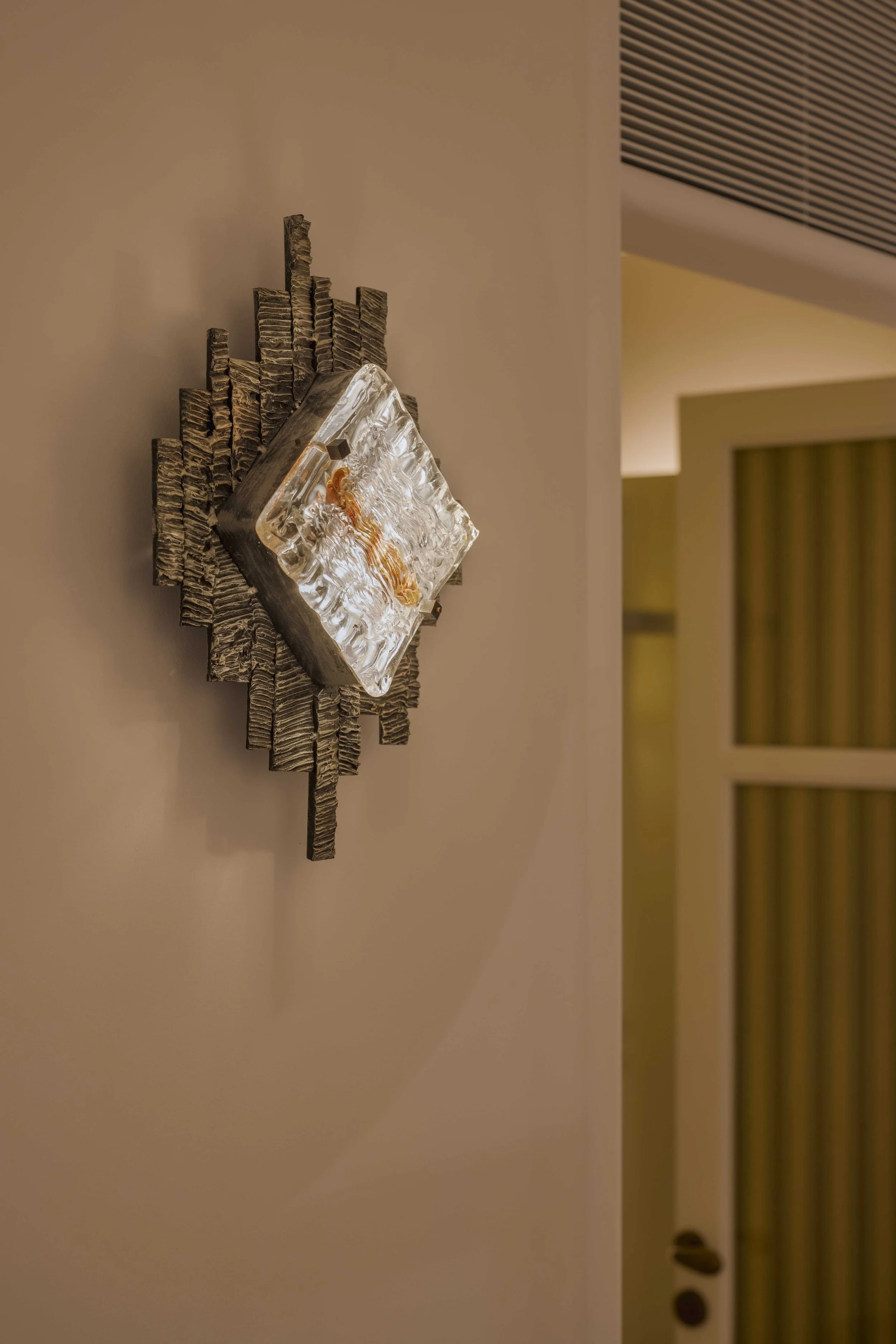 Close-up of an artistic wall sculpture with textured wooden pieces and a glass block with an abstract design, mounted on a beige wall in an indoor space.