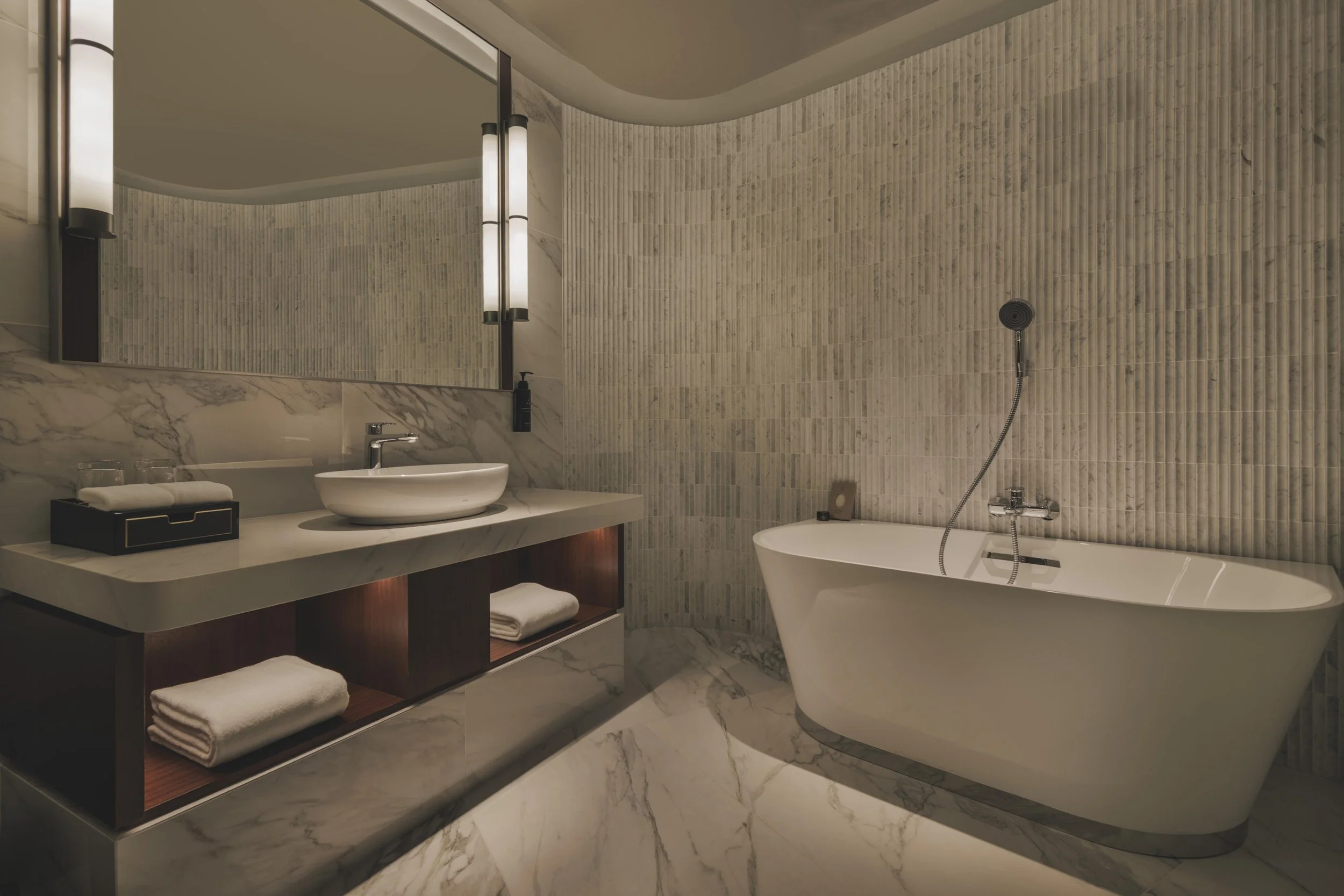 Modern bathroom with marble floor and walls, featuring a bathtub, vanity with sink, a large mirror, and wall-mounted lighting.