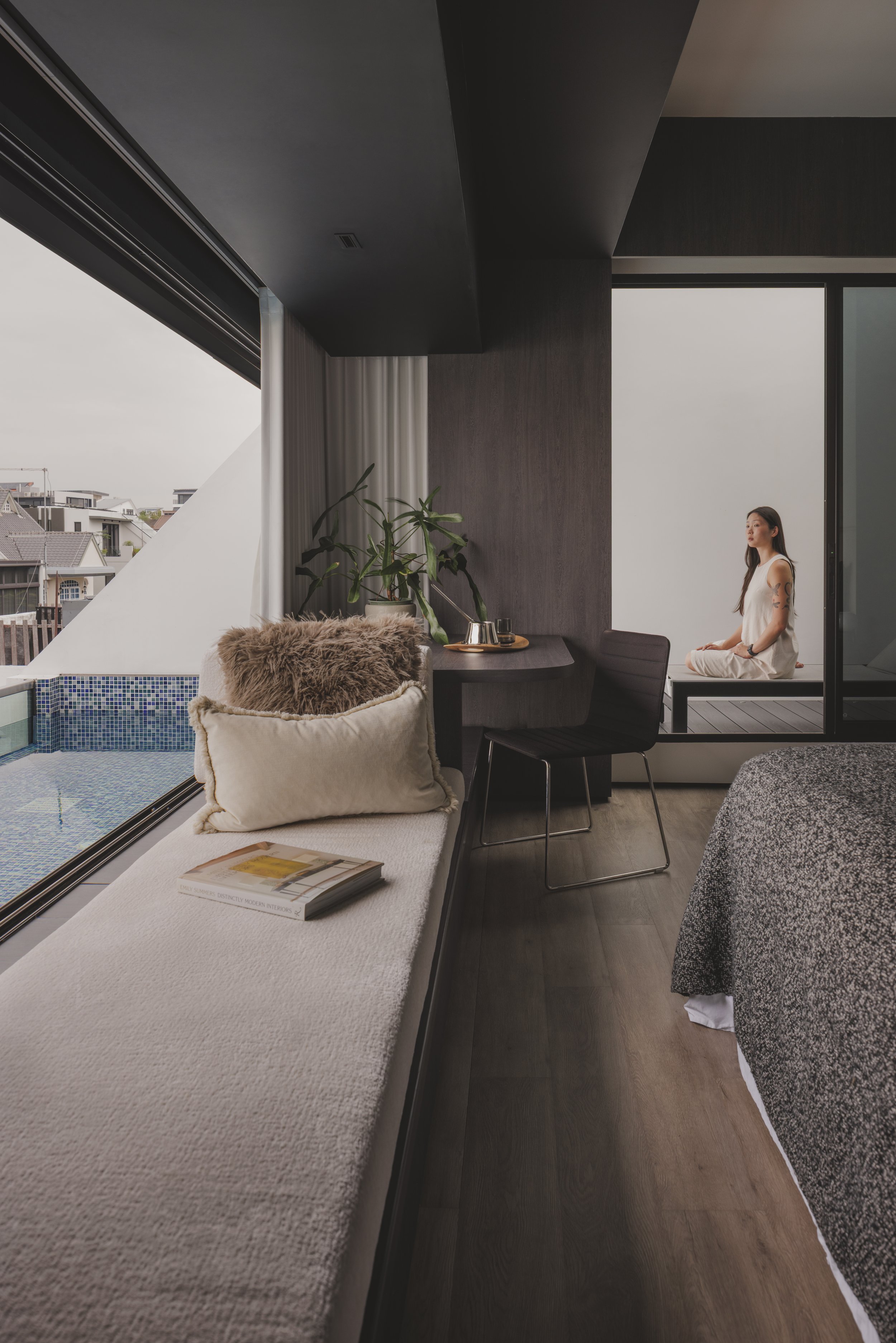 Interior of a modern living room with large windows, a white couch with a plush pillow and a magazine, dark wood flooring, and a woman practicing yoga or meditation near the window.
