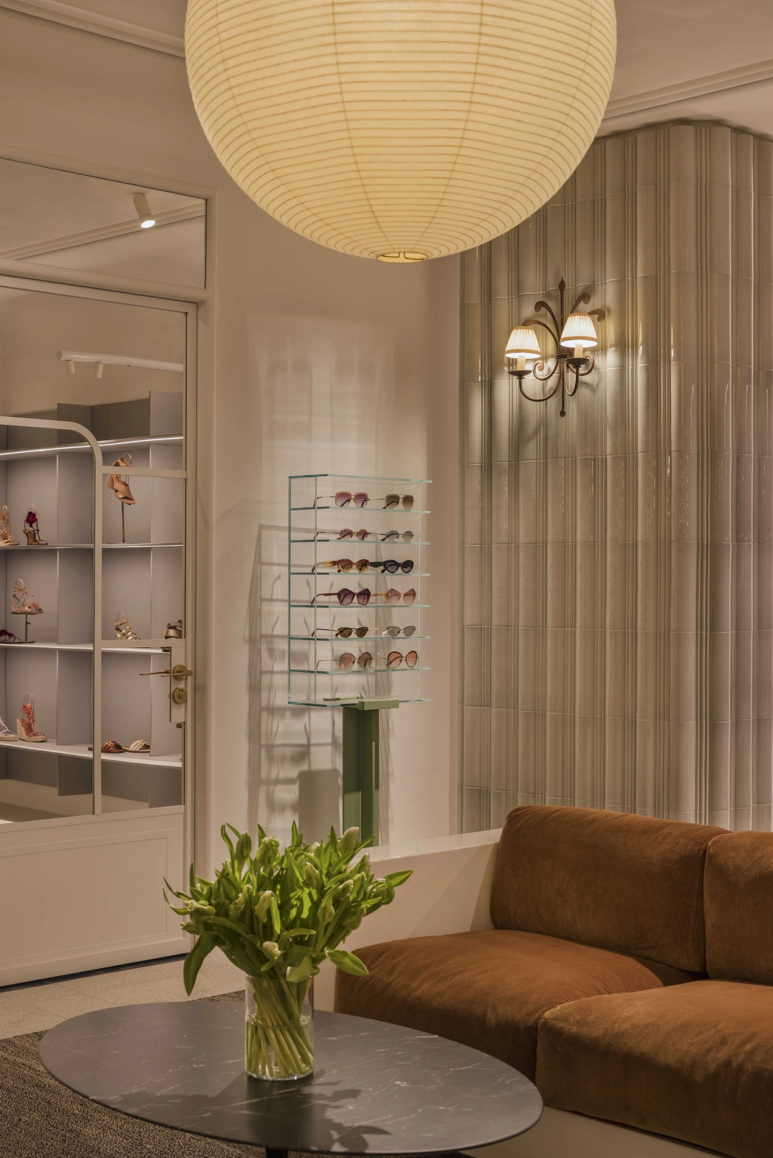 Interior of a shopping or retail store with display shelves of sunglasses and shoes, a black marble table with a clear glass vase of green plants, a brown couch, and a large paper lantern ceiling light.