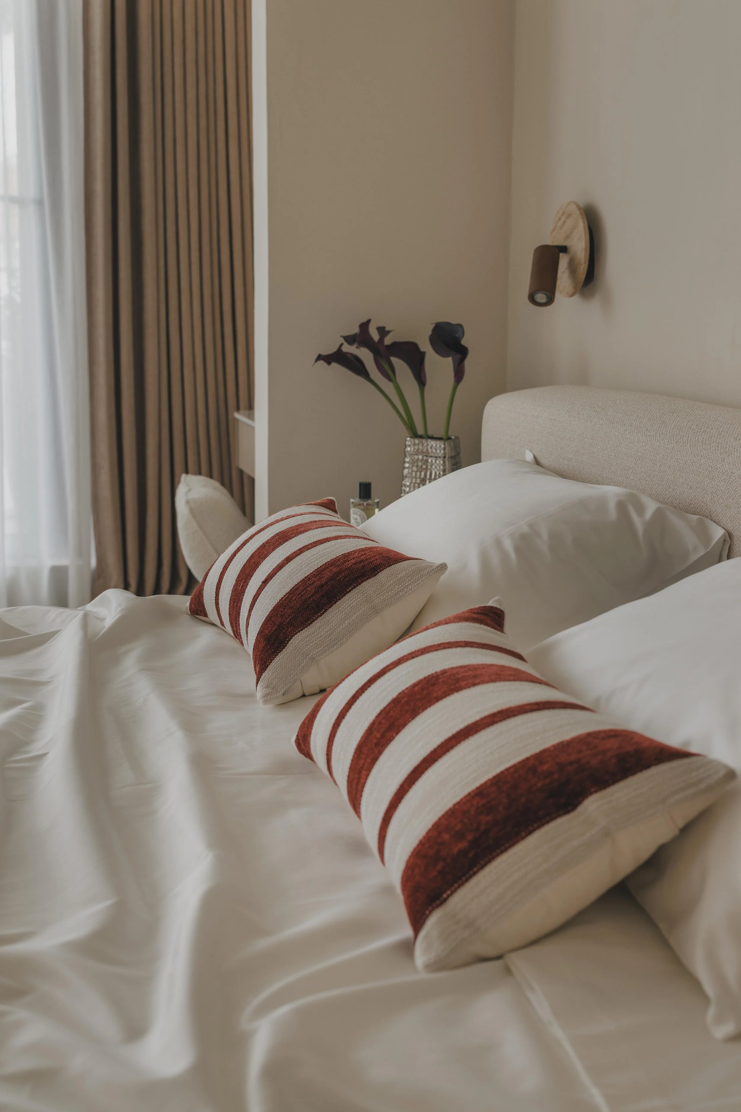 A bed with a white comforter and striped pillow, a potted plant with dark purple flowers on a nightstand, with beige walls and a wall-mounted light fixture.
