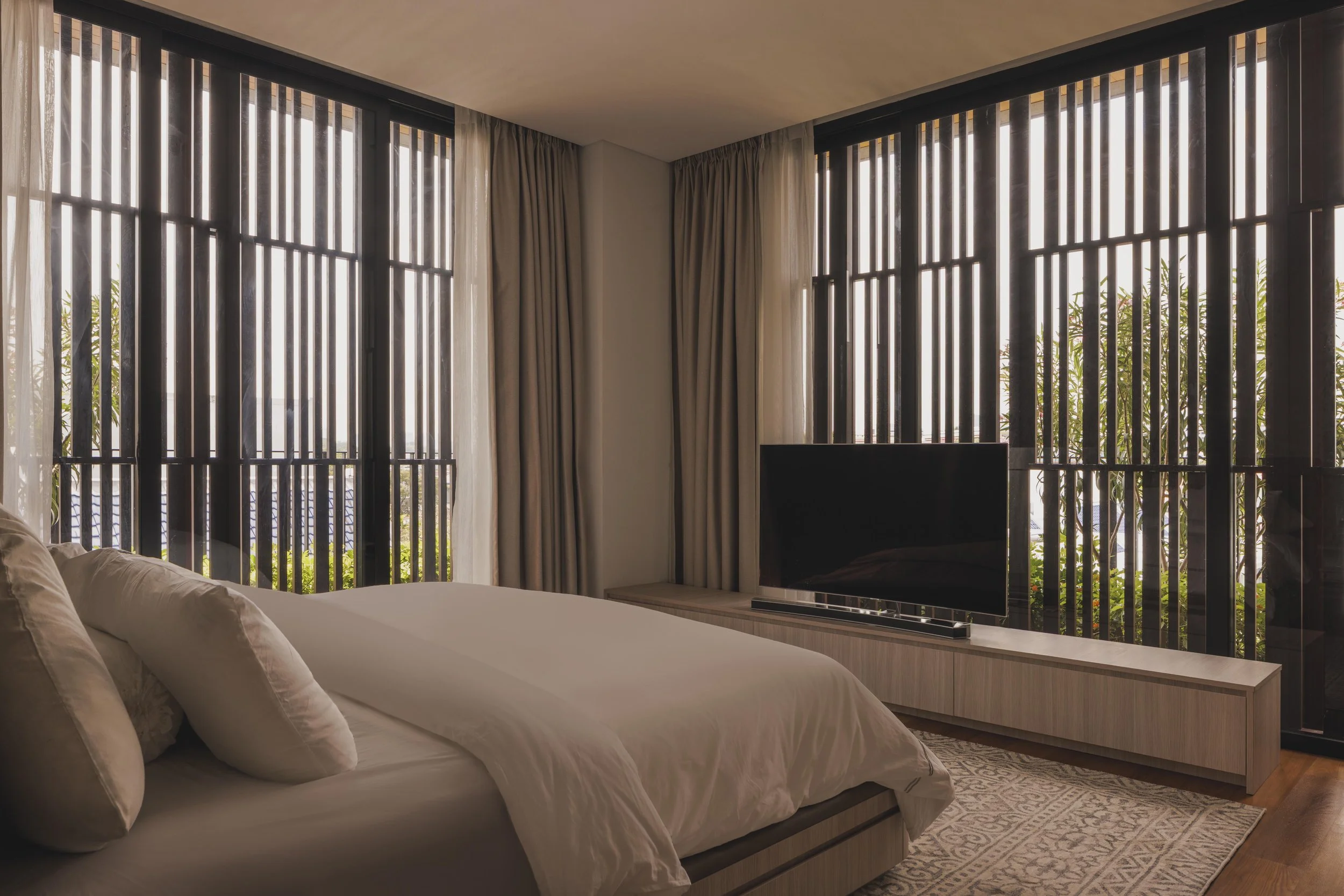 A bedroom with a bed and white pillows facing large windows with vertical wooden blinds and cream curtains, and a flat-screen TV on a wooden console beneath the windows.