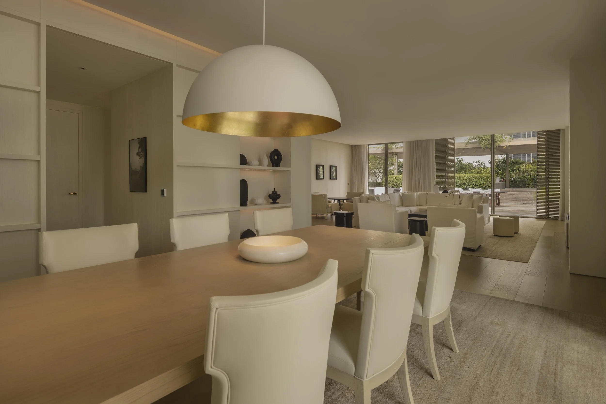 Modern open-concept living and dining area with a large wooden table, white chairs, a hanging dome pendant light with a gold interior, and a neutral color scheme. Large windows and sliding glass doors offer outdoor views.
