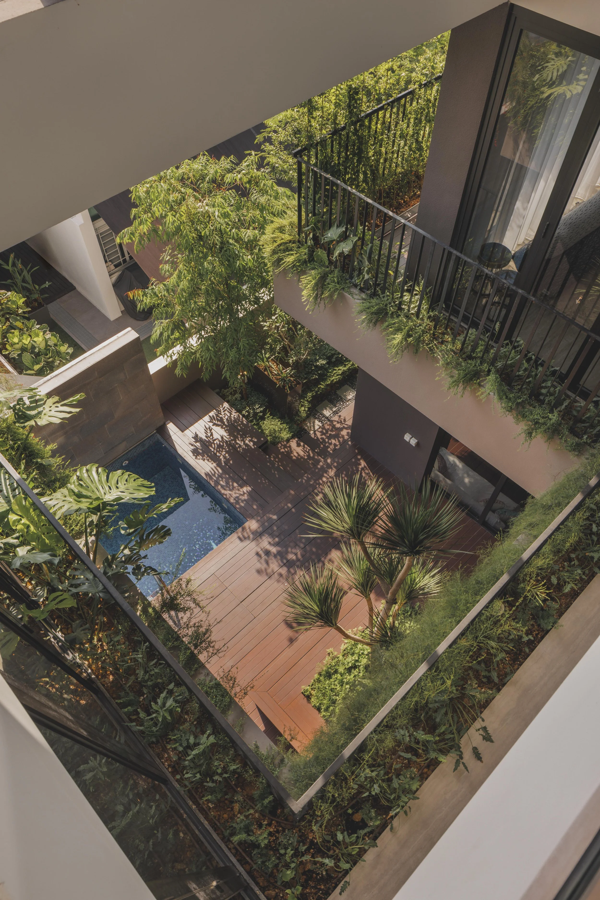 An aerial view of a high-rise apartment balcony filled with lush green plants and trees, overlooking a garden below on a sunny day.