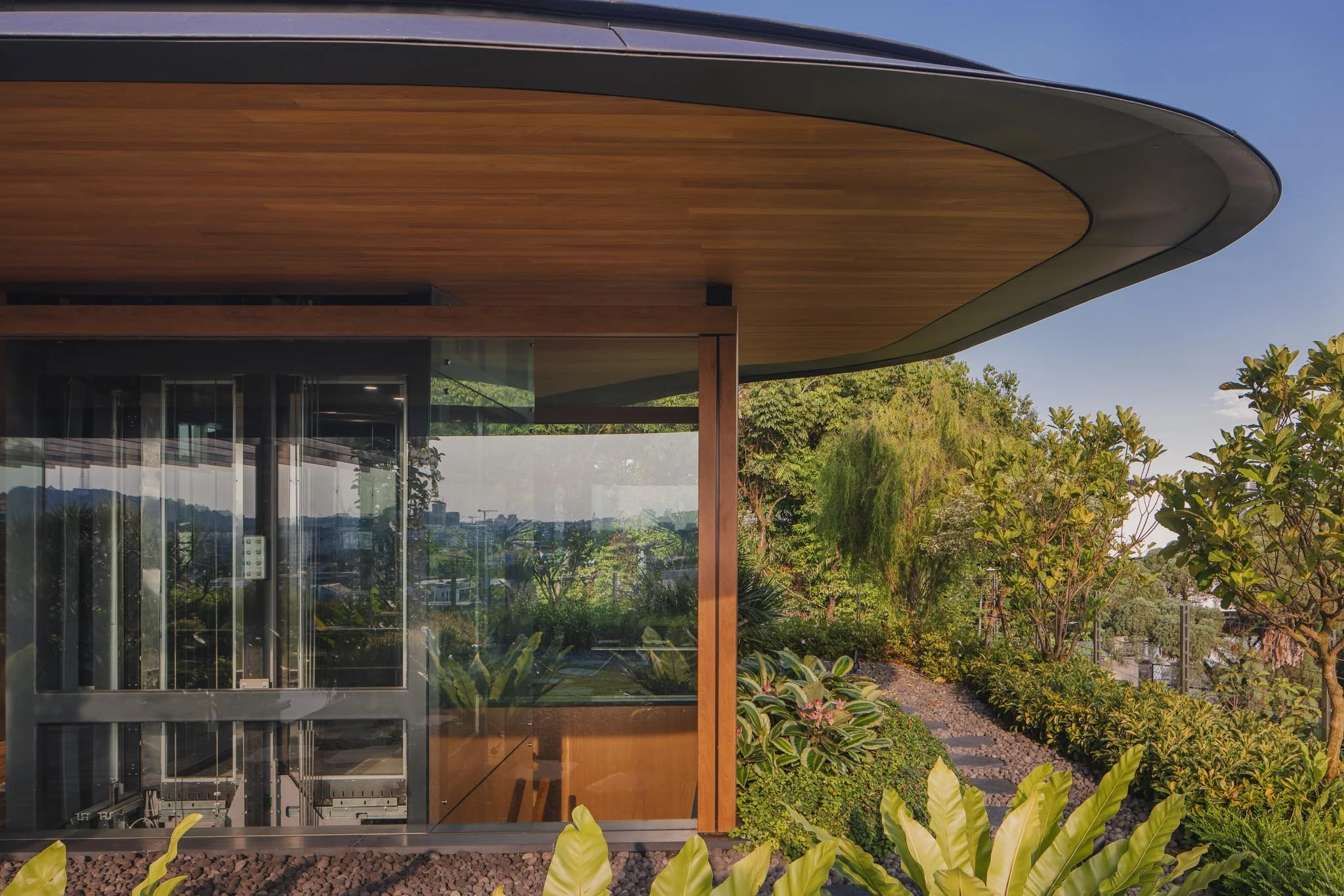 Modern building with curved roof, glass walls, and wooden accents, surrounded by green plants and trees, under a blue sky.