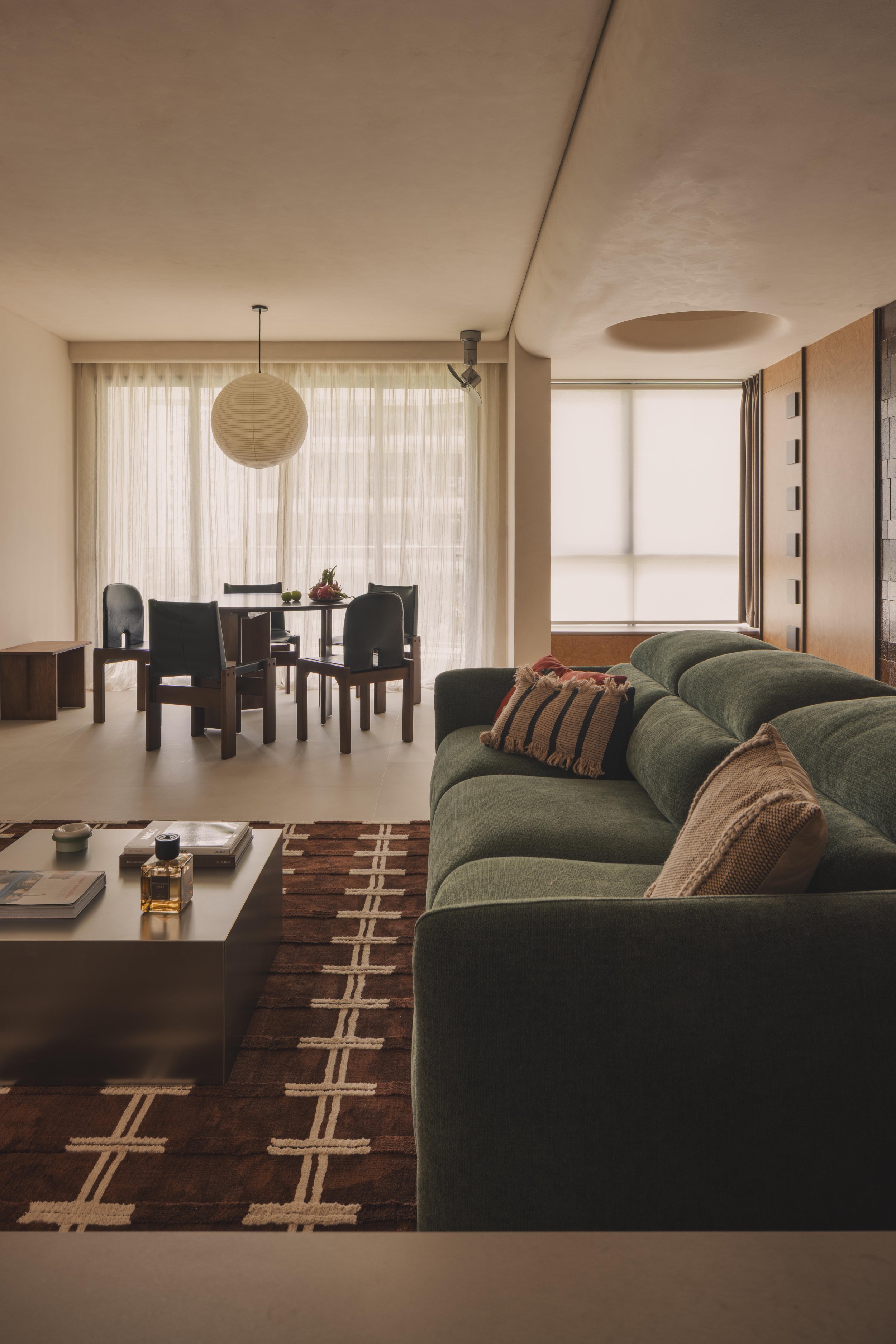 Living room with a green sofa, brown patterned rug, white coffee table with books and decorative items, a dining area with a round table and black chairs, and large windows with sheer curtains letting in natural light.