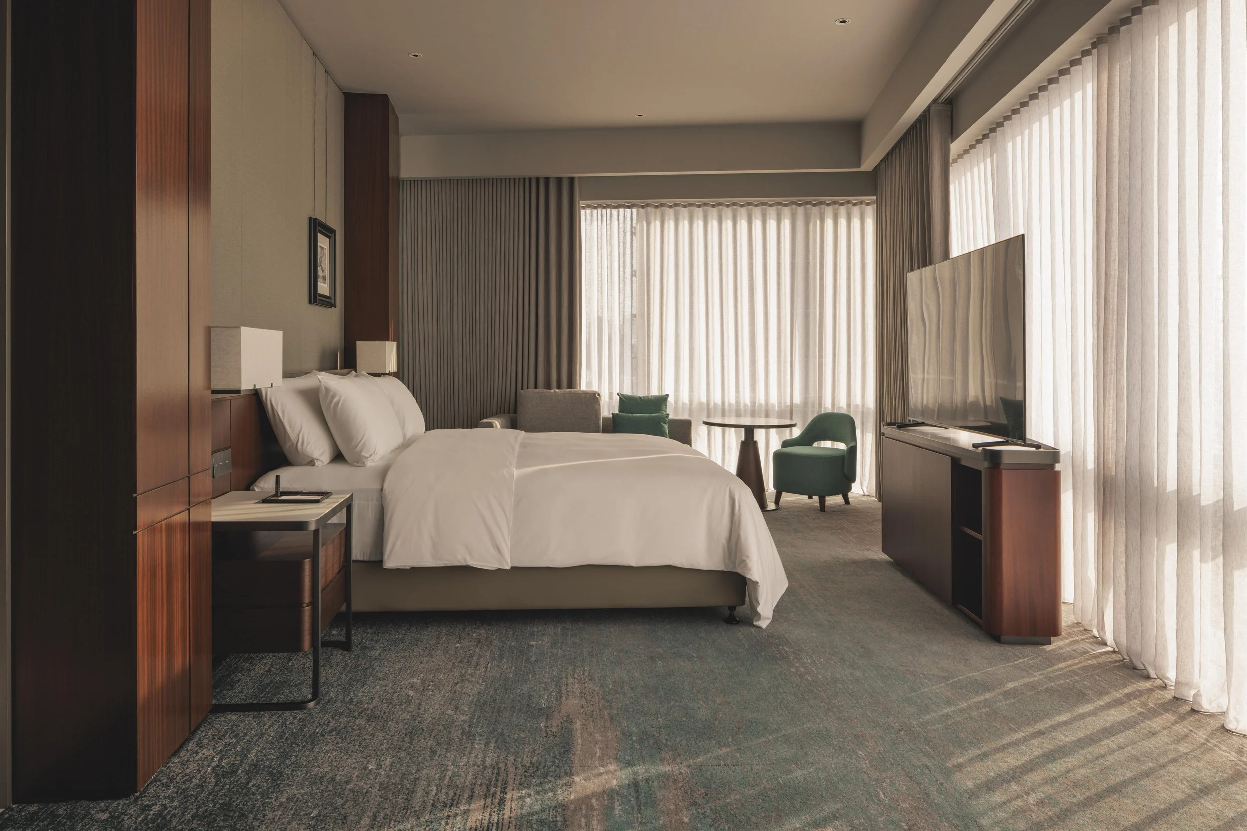 Hotel room with a bed, side table, armchair, and large windows with sheer curtains letting in natural light.