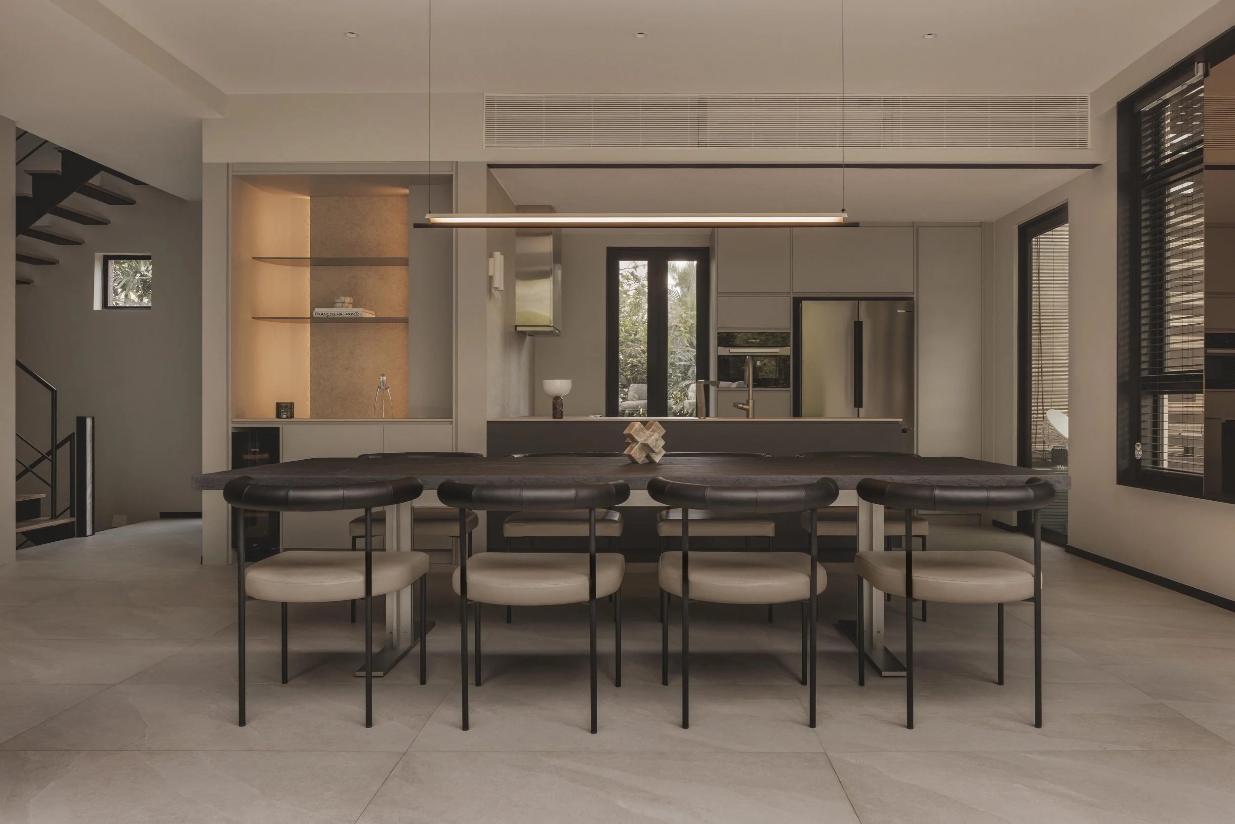 Modern kitchen with a large wooden dining table and upholstered chairs, cabinetry with built-in appliances, large windows with blinds, and minimalist decor.