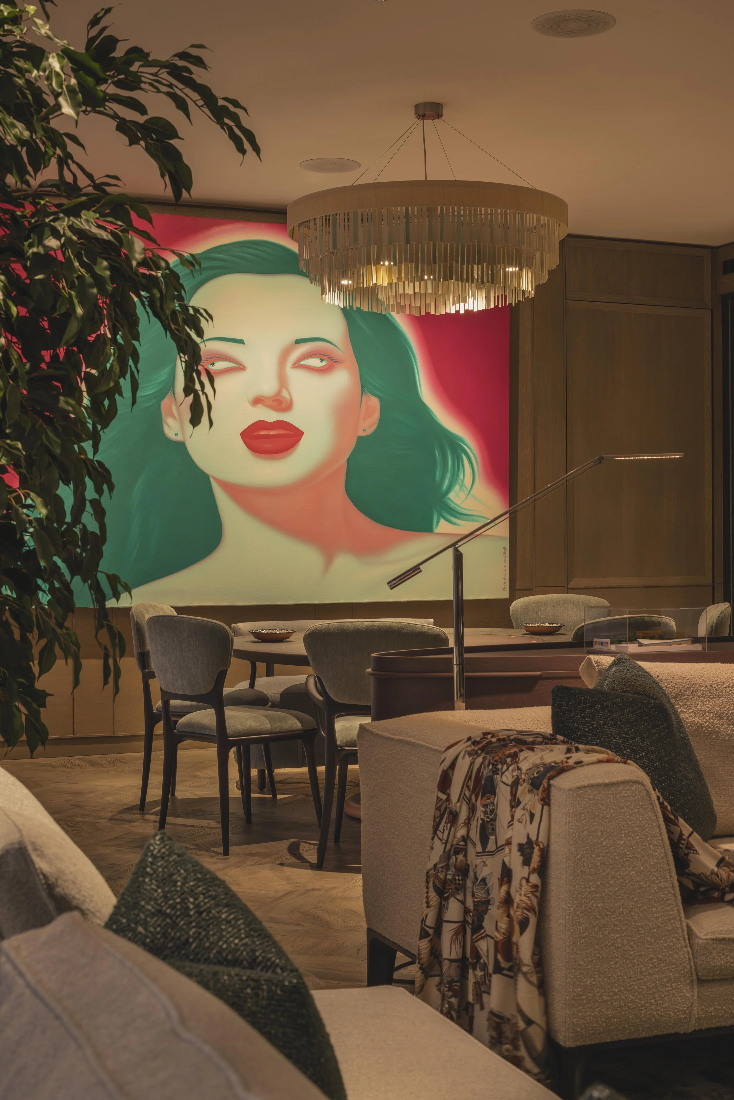A modern, stylish interior with a large vibrant portrait of a woman with green hair and red lips on the wall, surrounded by ceiling lights, a chandelier, a wooden wall, and various chairs and sofas with patterned cushions and throws.