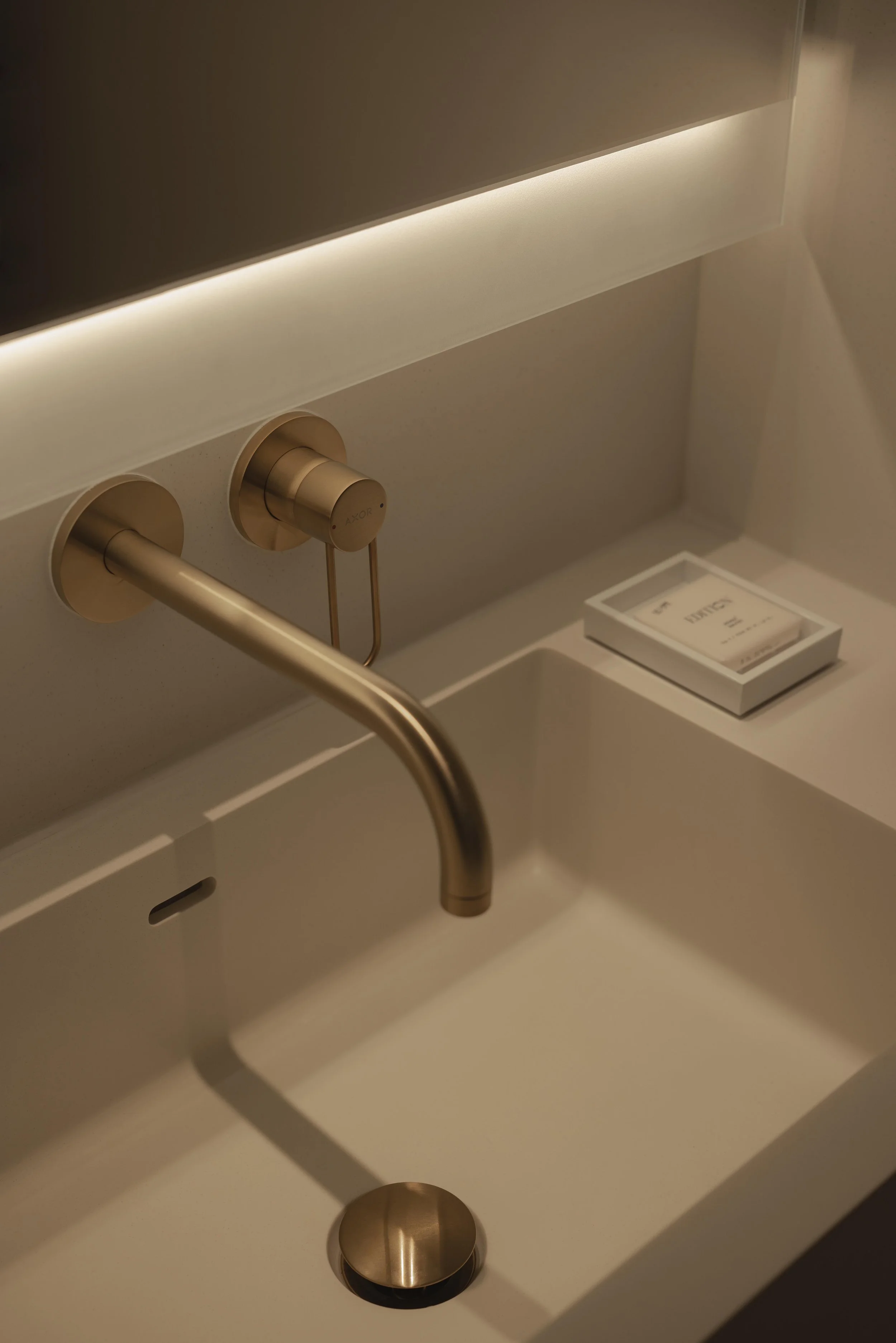Close-up of a modern sink with a gold faucet and a gold control knob, a white rectangular item inside a small box, and a wooden round object, all set against a beige wall and beige countertop.