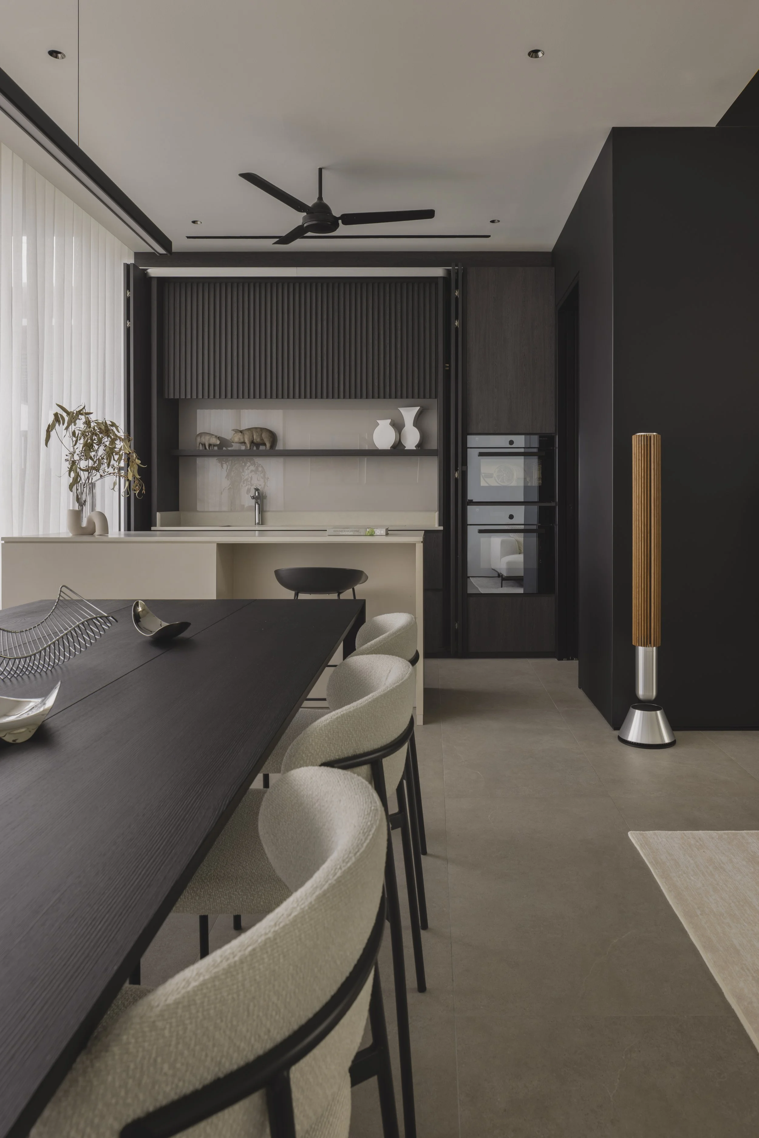 Modern dining area with beige chairs around a black table, decorative objects on the table, a plant, and a corner with a black wall, a speaker, and an artistic wooden lamp.