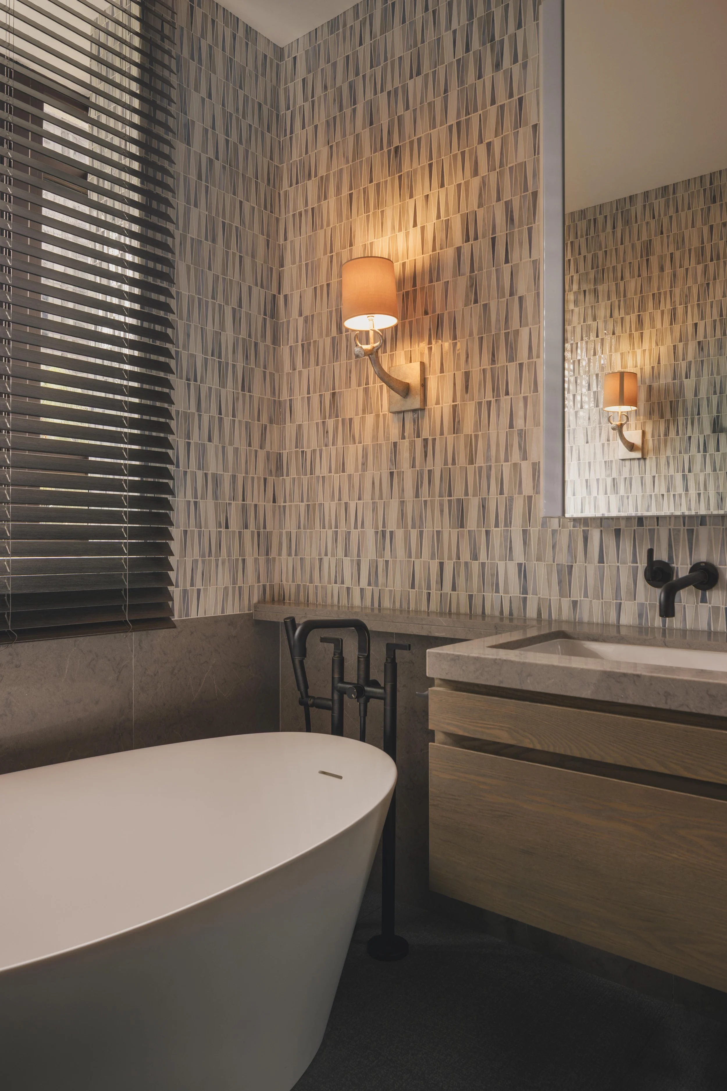 A modern bathroom features a freestanding white bathtub, a wooden vanity with horizontal drawers, and a textured geometric tile wall. A wall-mounted lamp with a beige shade provides warm lighting, and there's a large mirror reflecting the bathroom interior. The window has black horizontal blinds, and a black faucet is visible at the sink.