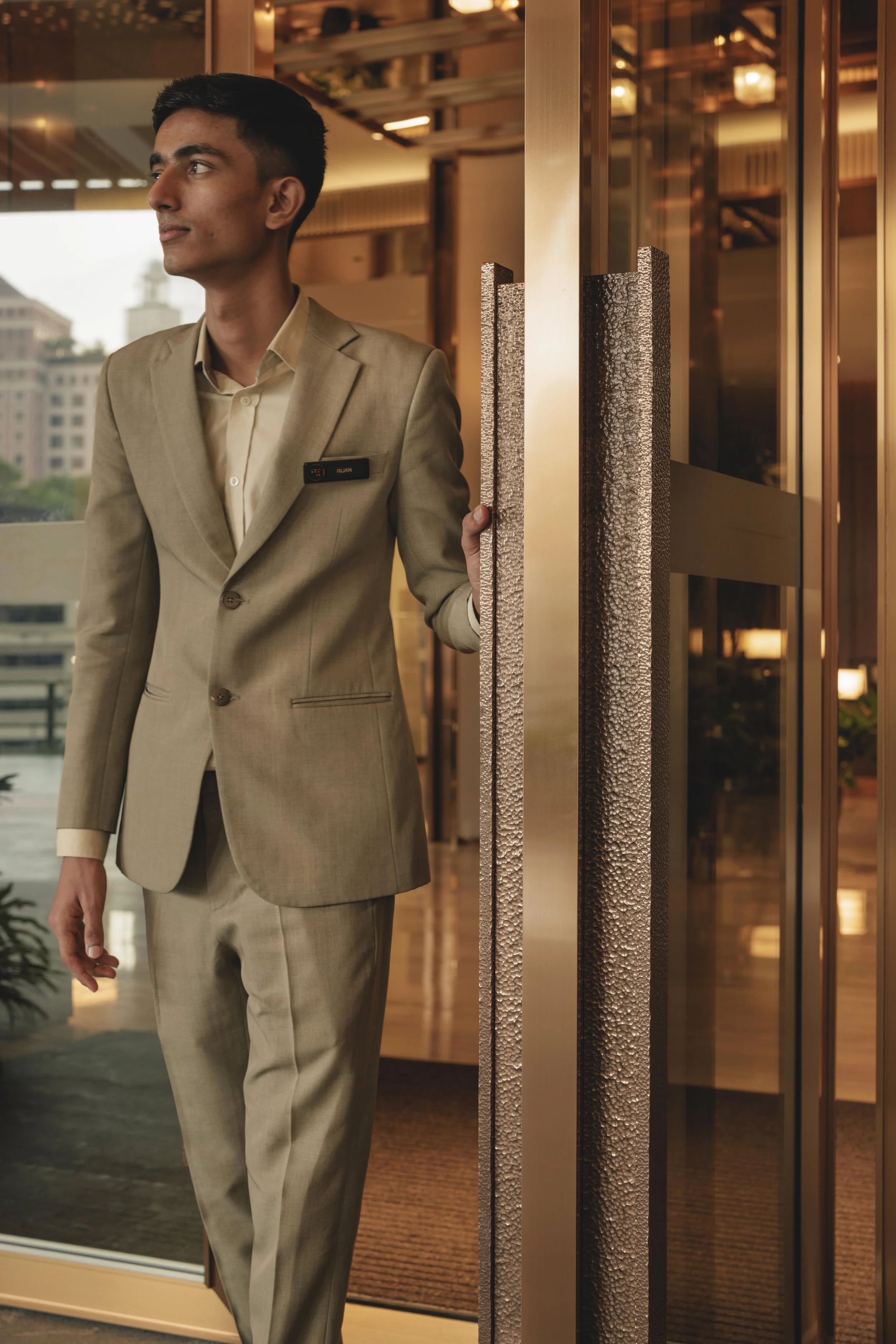 A man in a beige suit standing near glass doors inside a building with warm lighting and a city view in the background.