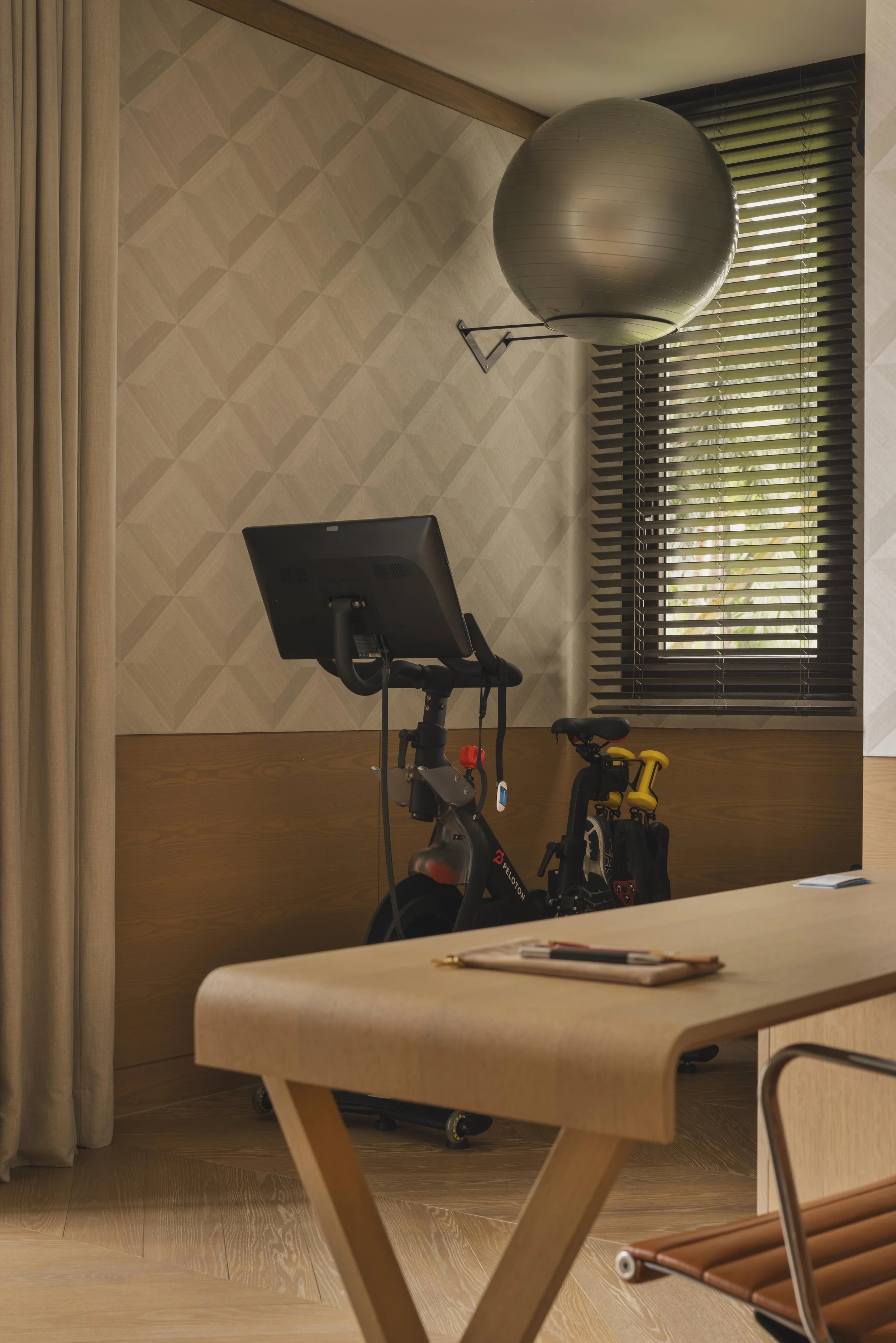 A room with a wooden exercise bike and children’s riding toys, a beige table in the foreground, a window with blinds, a wall with geometric pattern, and a large exercise ball mounted on the wall.