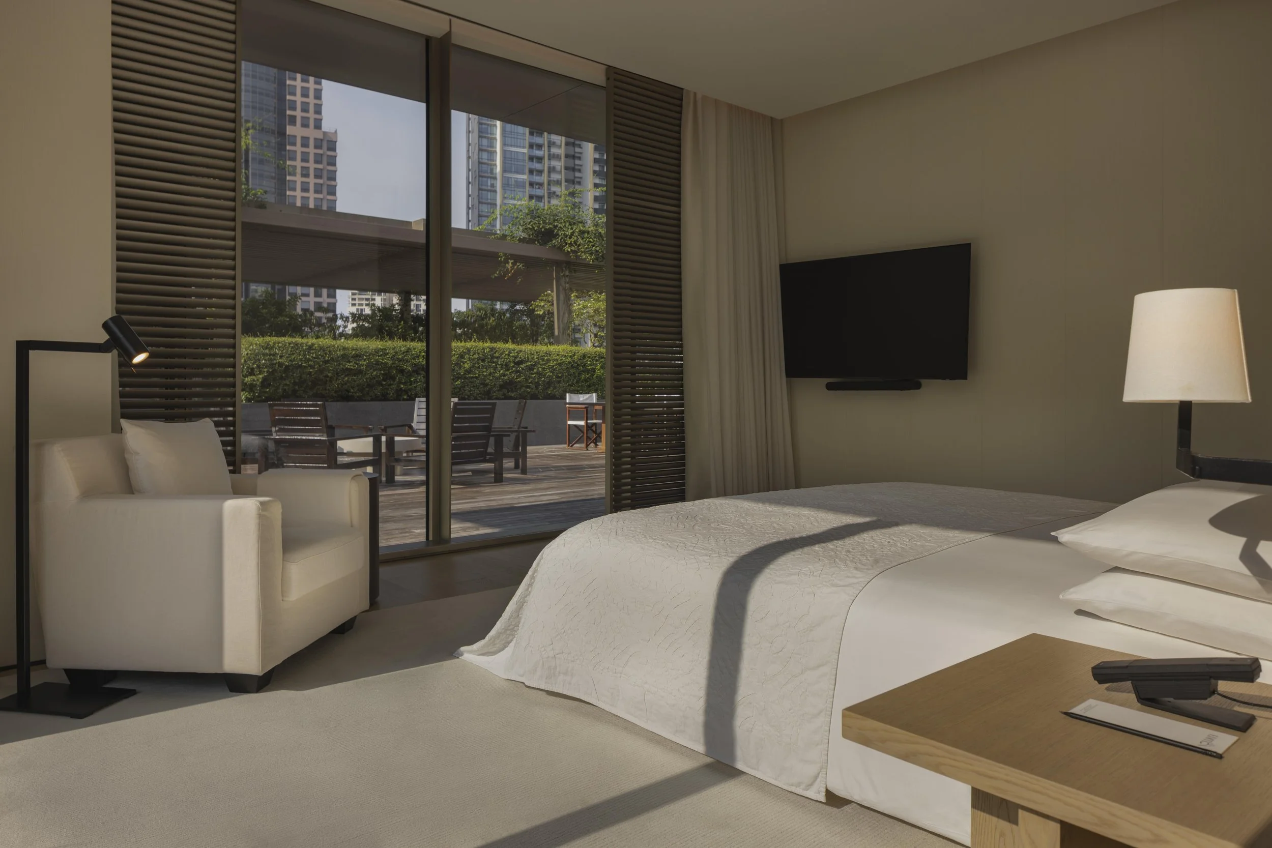 A modern hotel room with a large bed, a flat-screen TV mounted on the wall, a lamp, a small wooden table, and a sliding glass door leading to an outdoor balcony with chairs. The balcony overlooks tall city buildings, a hedge, and trees.