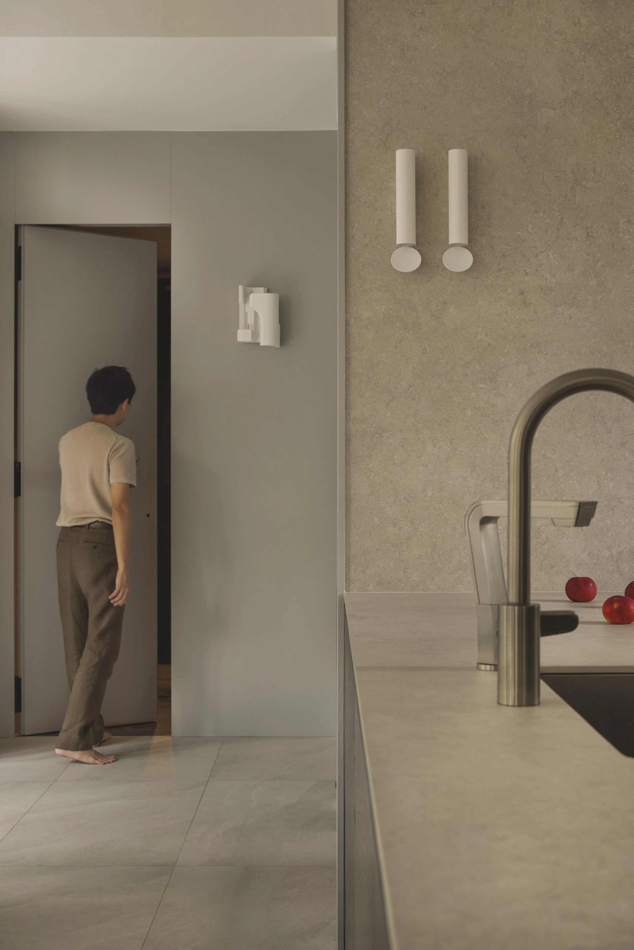 A person with short dark hair, wearing a beige t-shirt and brown pants, standing barefoot in a modern kitchen, with a gray door slightly open behind them. The kitchen has a beige countertop with red apples, a stainless steel faucet, and a light-colored backsplash.