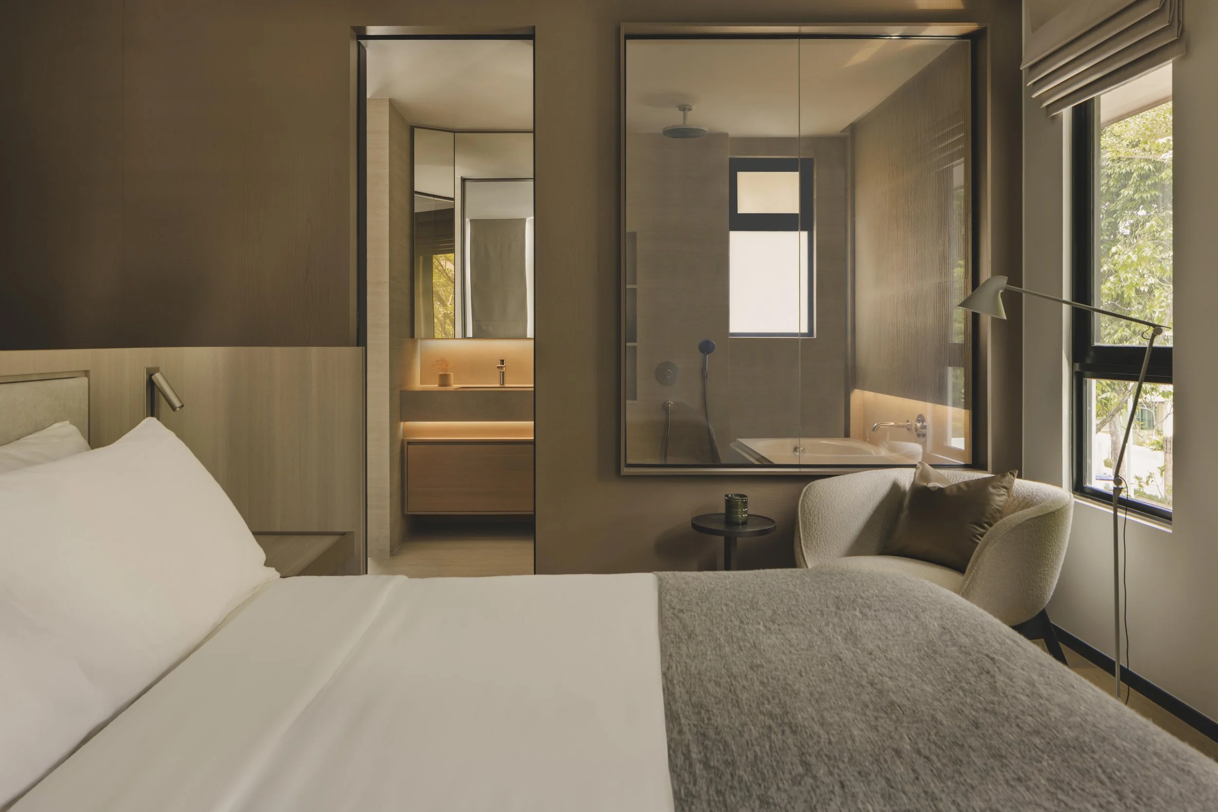 A modern hotel room with a bed, a sitting area with an armchair, a small side table with a candle, and a glass-enclosed bathroom with a bathtub, sink, and large windows showing outside greenery.
