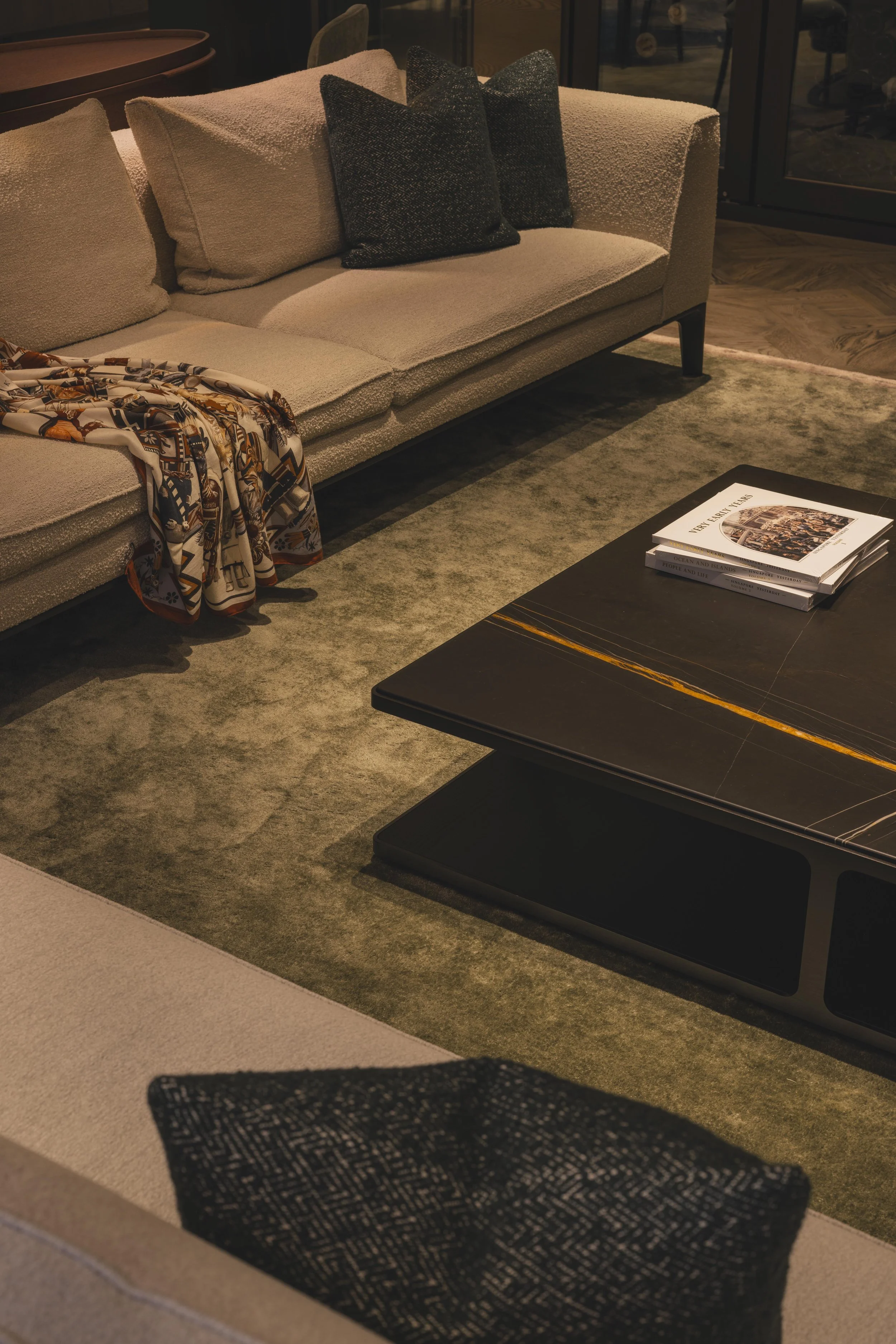 A beige sofa with black and beige cushions, a rolled-up patterned scarf hanging over the arm, and a black coffee table with magazines on it in a cozy living room.