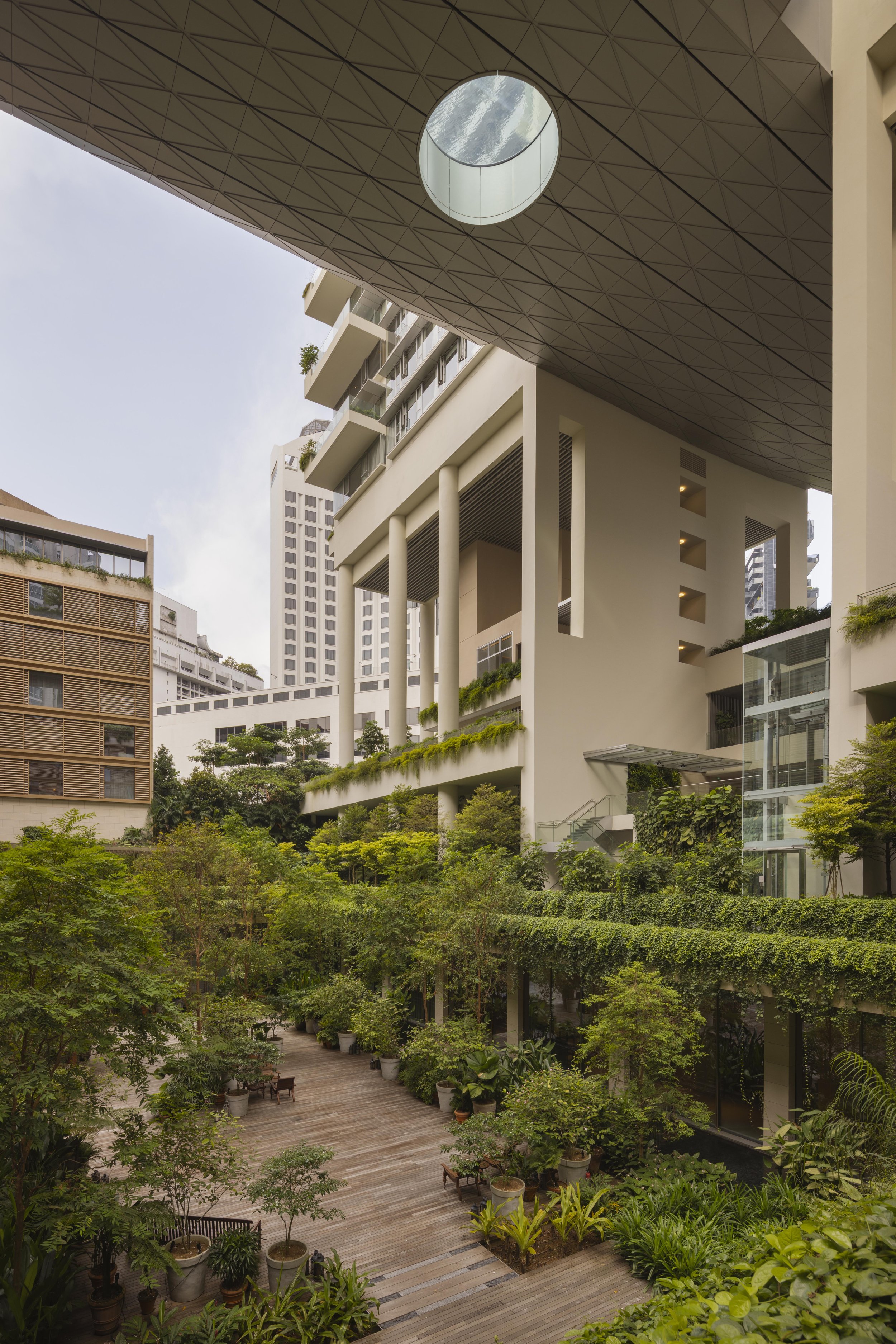 A modern building with a large circular opening in the roof, surrounded by greenery, trees, and potted plants on a wooden patio area.