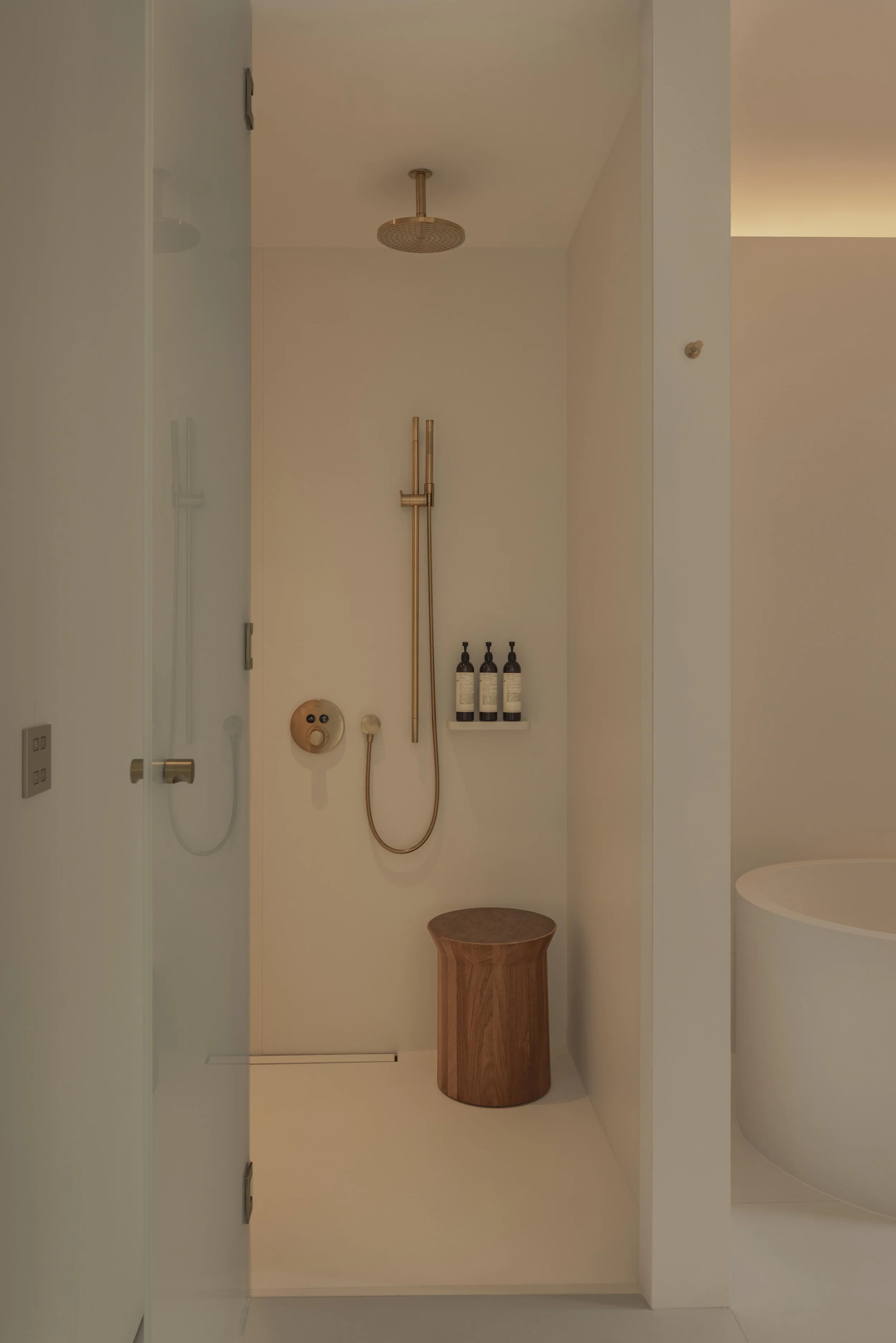 Minimalist bathroom with a walk-in shower, gold fixtures, three black bottles on a shelf, a round wooden stool, and a white bathtub.