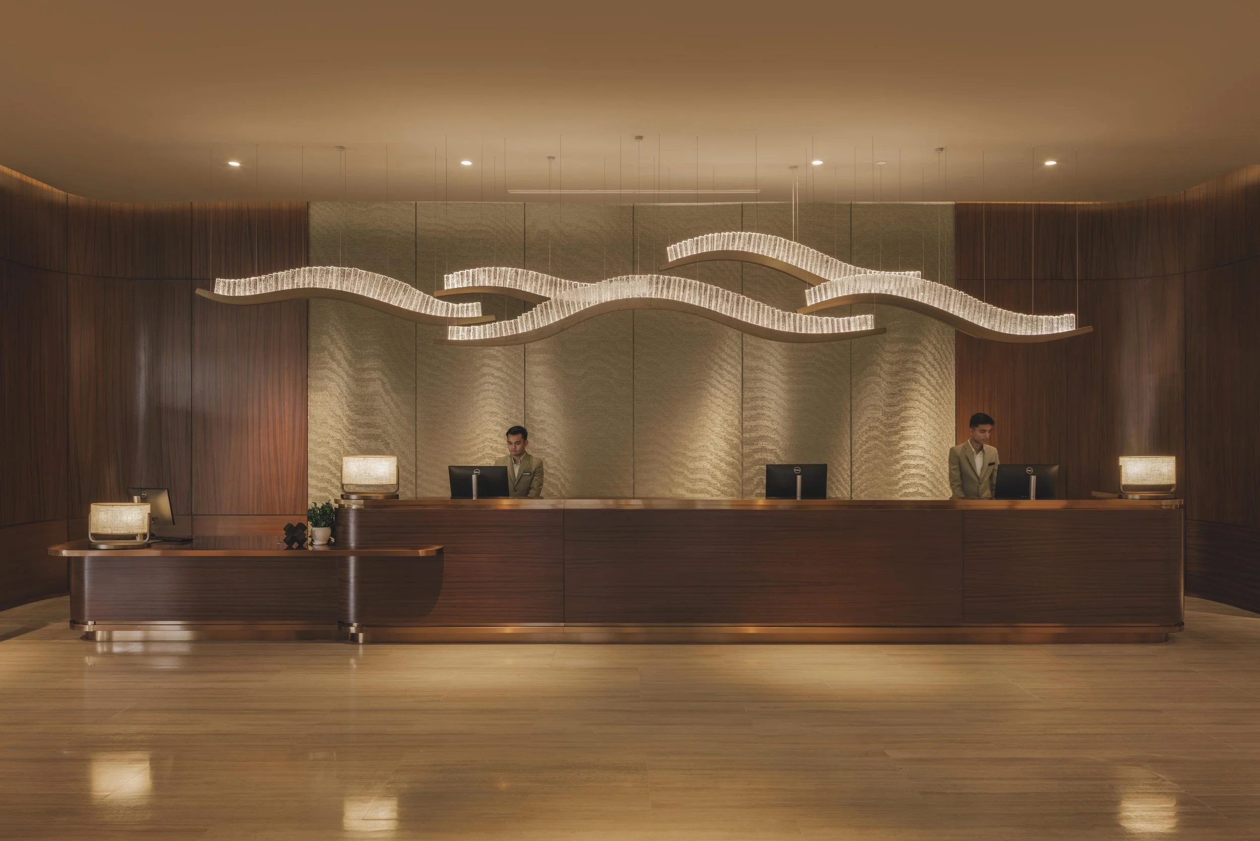 A hotel reception desk with a wavy modern chandelier overhead. Two staff members in suits stand behind the desk.