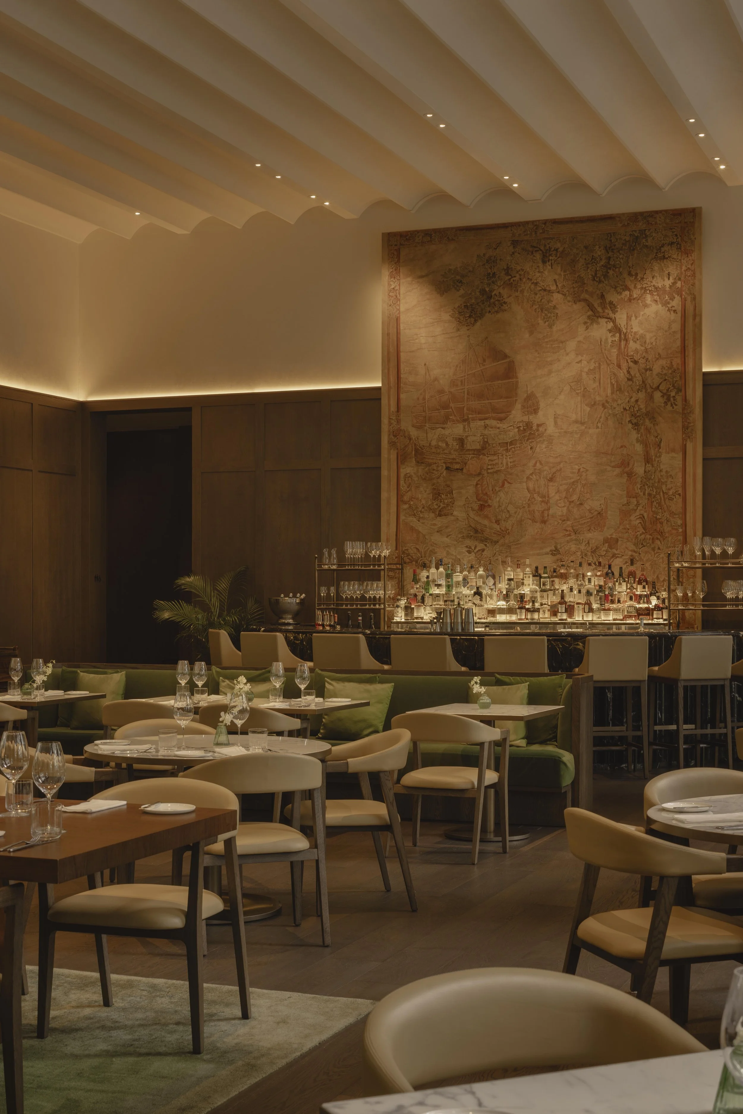 A sophisticated restaurant interior with wooden paneling, a large tapestry featuring a sailing ship, and a bar area with liquor bottles. Dining tables are set with glasses and napkins, and light creates a warm ambiance.