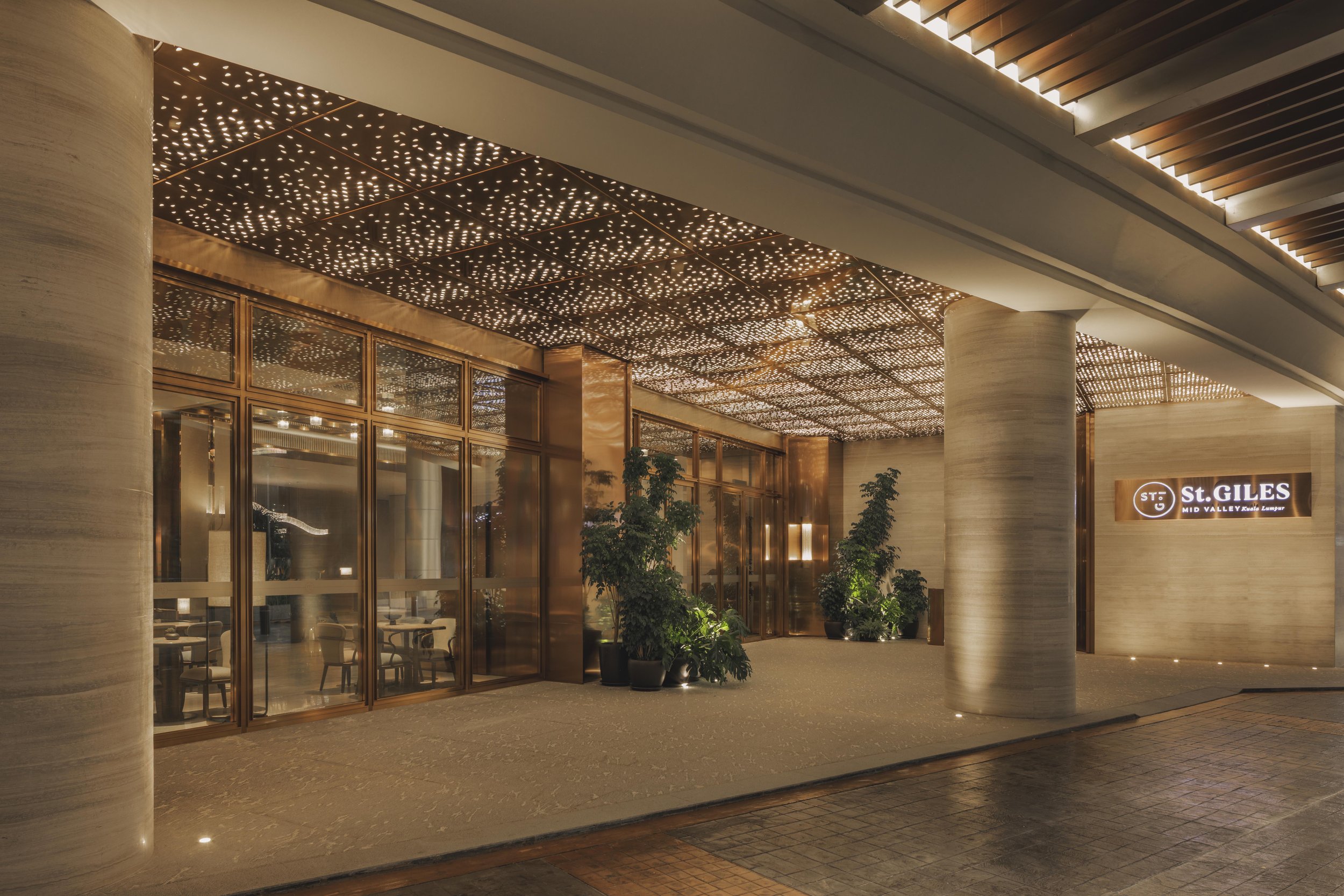 The image shows the entrance of a hotel named St. GILES, located in Mid Valley, Kuala Lumpur. The area is well-lit with a textured ceiling featuring perforated panels with small circular lights, and large glass doors framed in a metallic rose gold color. There are potted plants near the entrance, and the overall ambiance is warm and inviting.