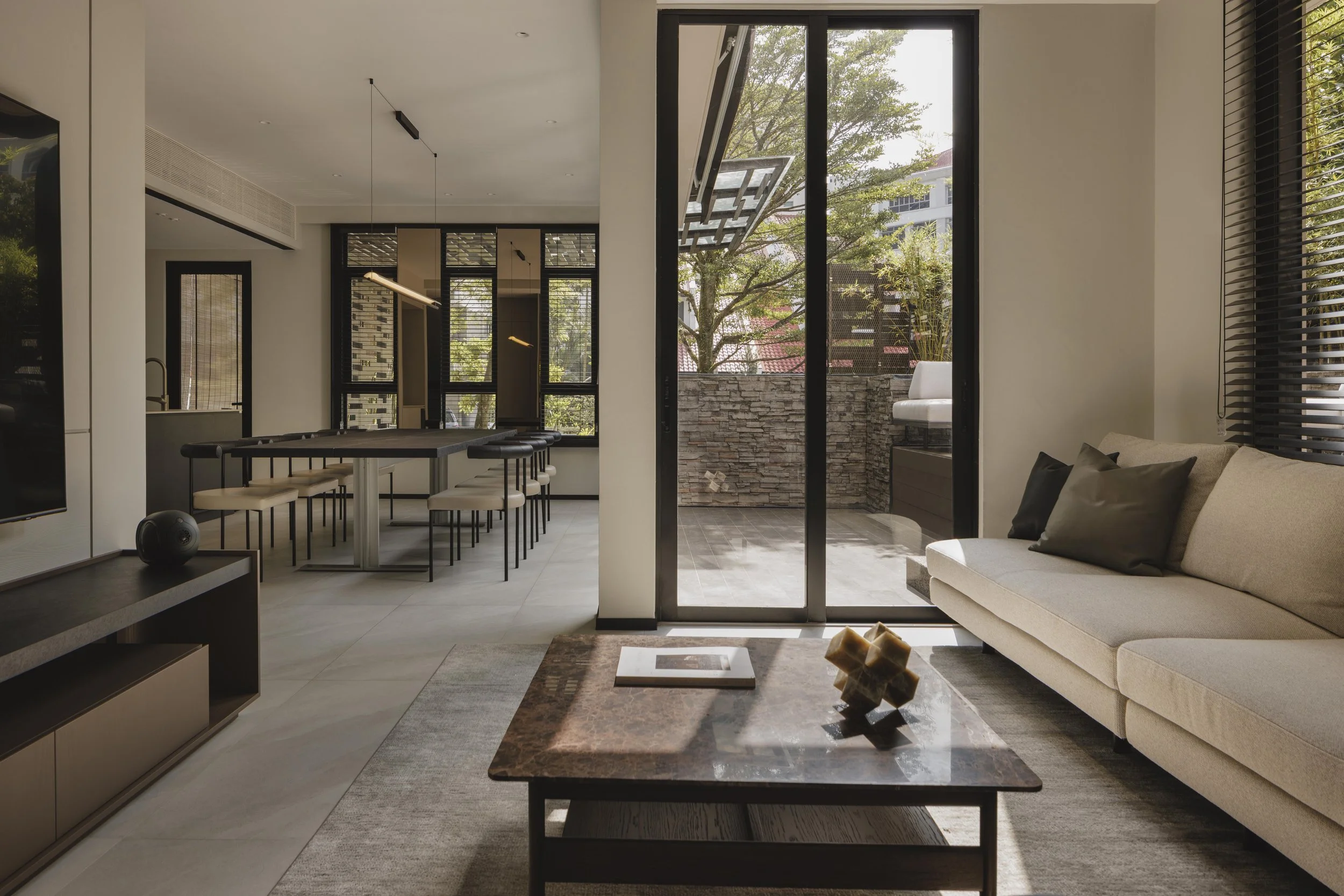 Modern living room with a beige sofa, black shutter blinds, and large glass sliding doors opening to a patio with trees and a stone wall.