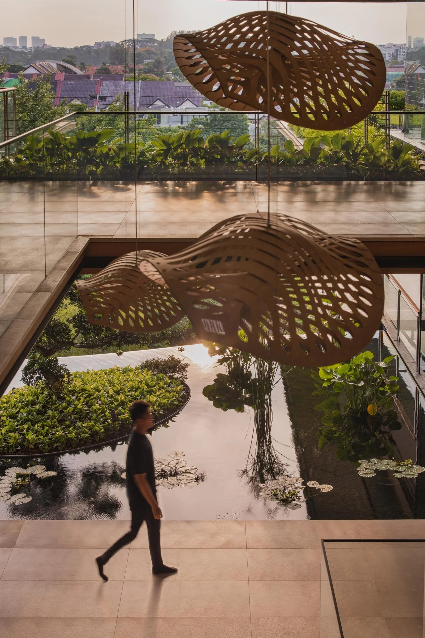 A person walking past a pond with water lilies inside a modern building with artistic wooden ceiling lamps and a scenic view of a city skyline.
