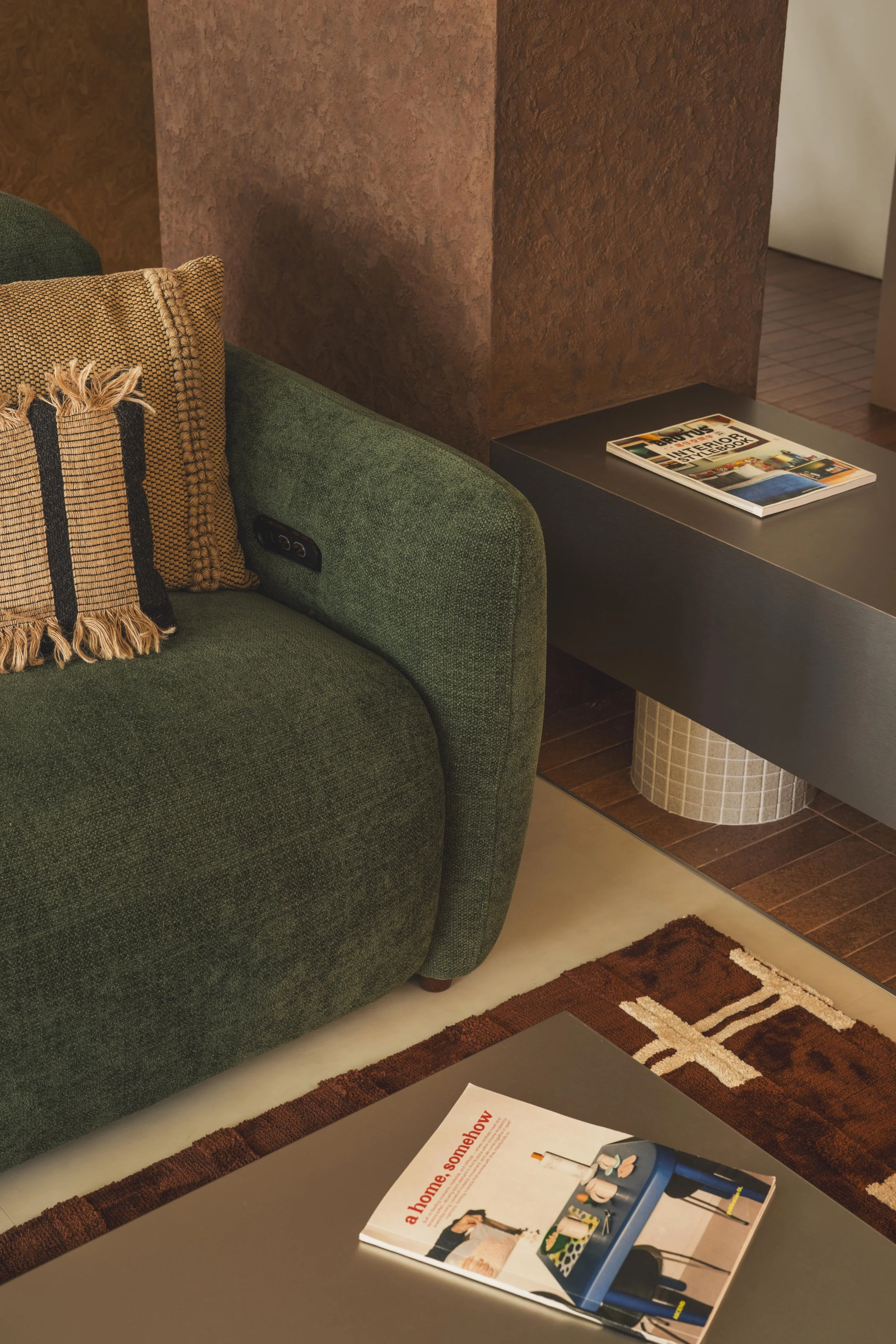 Close-up of a green upholstered sofa with a tan and black patterned pillow, next to a black table with a magazine on it. Part of a textured brown wall and a portion of a patterned brown and beige rug are visible.