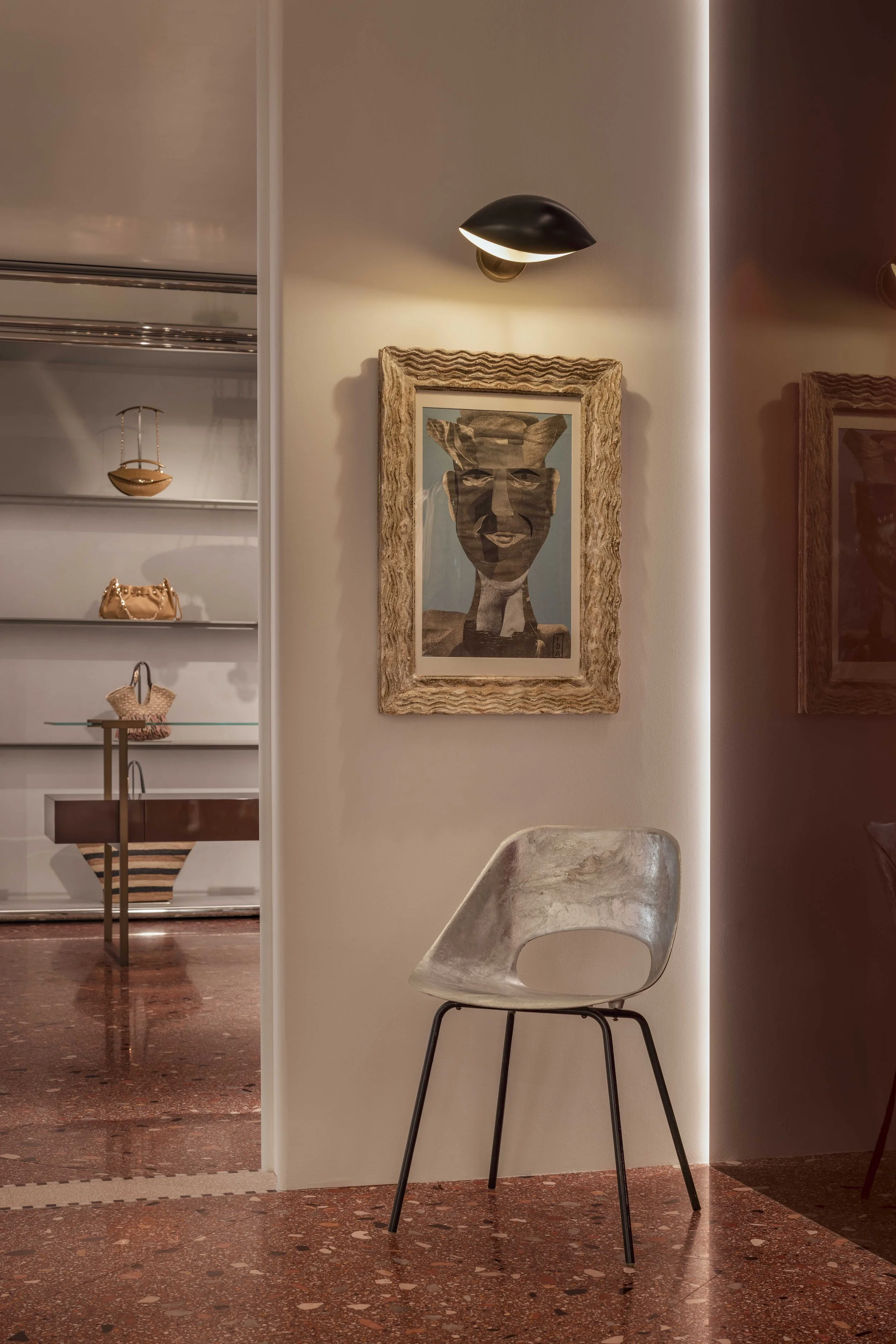 Interior of a modern room with a metallic chair, a artwork of a face with a large head framed in rustic wood, a wall-mounted black light, and a wall with built-in shelves displaying handbags and decor. The floor is polished reddish-brown with terrazzo pattern.