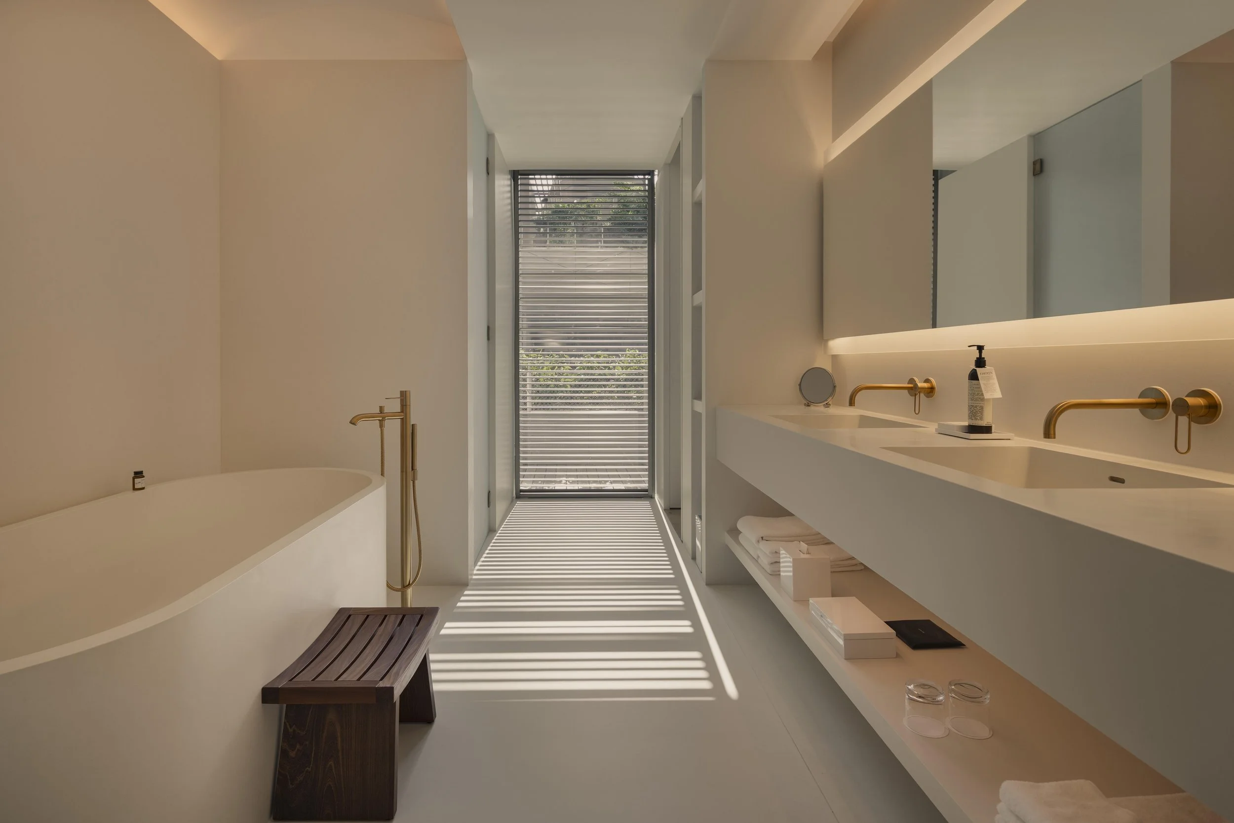 Modern bathroom with white walls, a built-in white sink with brass fixtures, a large mirror with backlighting, a wooden bench in the shower area, and a glass door with horizontal blinds leading outside, letting in natural light.
