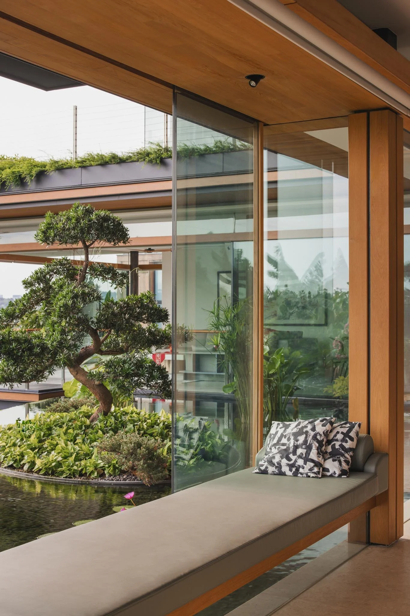 An indoor lounge area with a glass wall overlooking a garden pond filled with lush greenery and a tree. There is a cushioned bench outside the window with a decorative pillow, and the ceiling is made of wood.